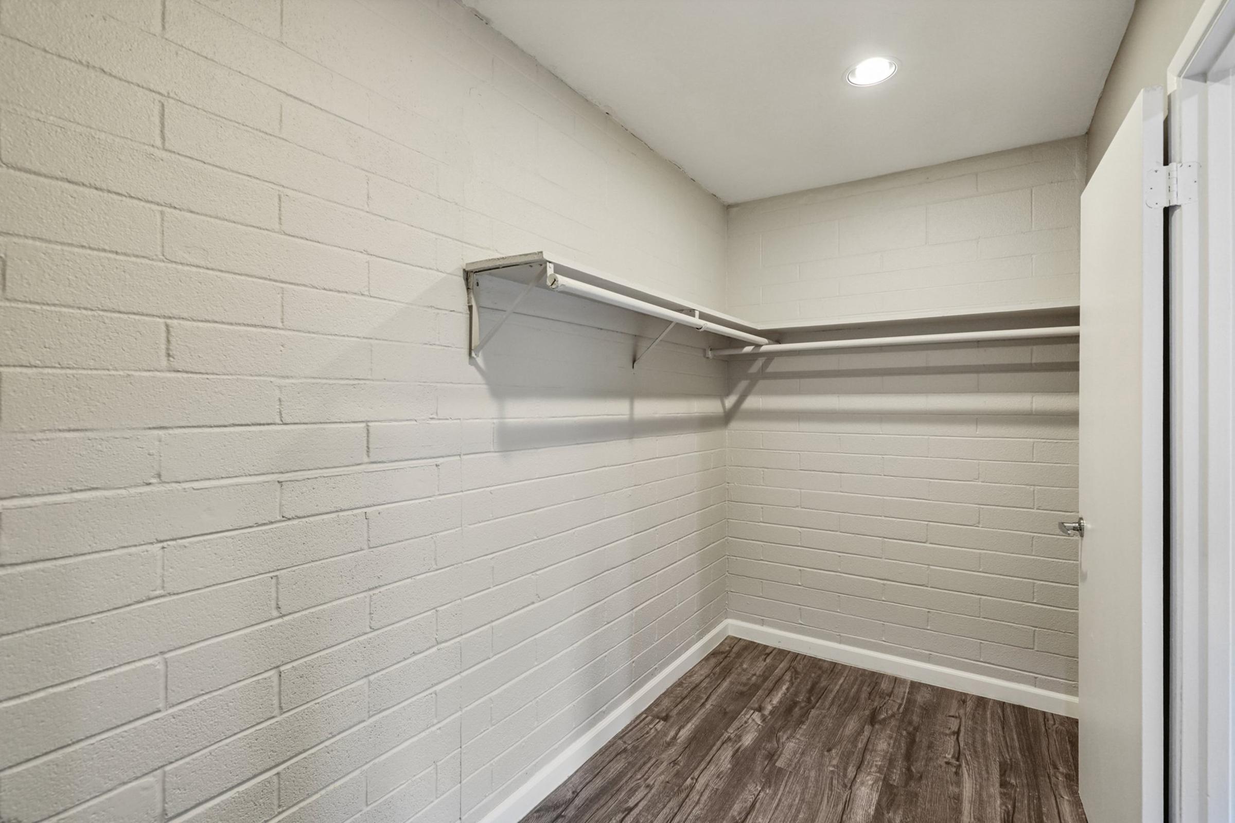 Empty closet with a light-colored painted concrete wall, a single built-in shelf along one side, and a wooden floor. The space is well-lit, featuring a ceiling light fixture and a white door partially visible on the right.