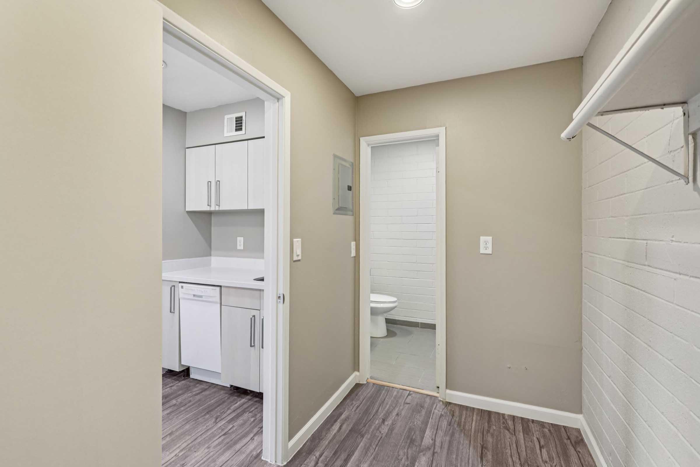 A small, modern utility area featuring a doorway leading to a bathroom on the right and a kitchen-like space with white cabinets and appliances on the left. The room has light gray walls and a wooden floor, providing a clean and functional layout. Natural light is present through overhead lighting.
