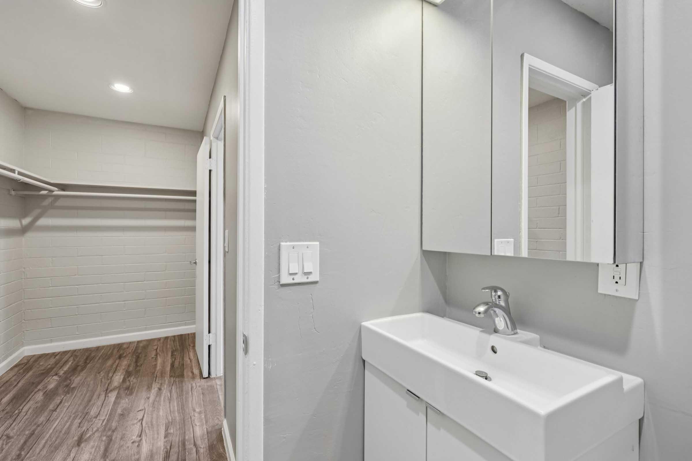 A modern bathroom featuring a wall-mounted sink with a faucet, a large mirror above it, and neutral gray walls. In the background, there is an open closet with shelving and wooden flooring, creating a clean and minimalist aesthetic.