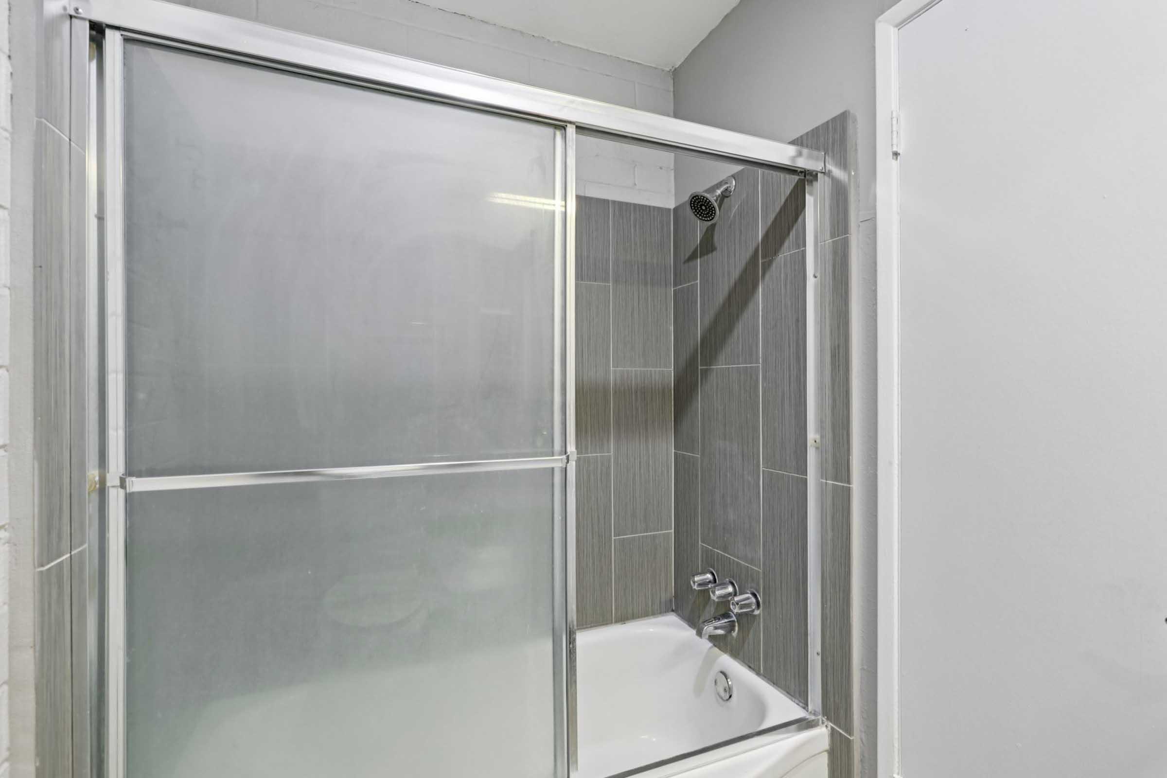 A modern bathroom featuring a glass shower enclosure with sliding doors. The shower has gray tiles on the walls and a showerhead above. A white bathtub is visible at the bottom, and there is a plain door on the right side of the image. The walls are painted light gray, creating a clean and contemporary look.