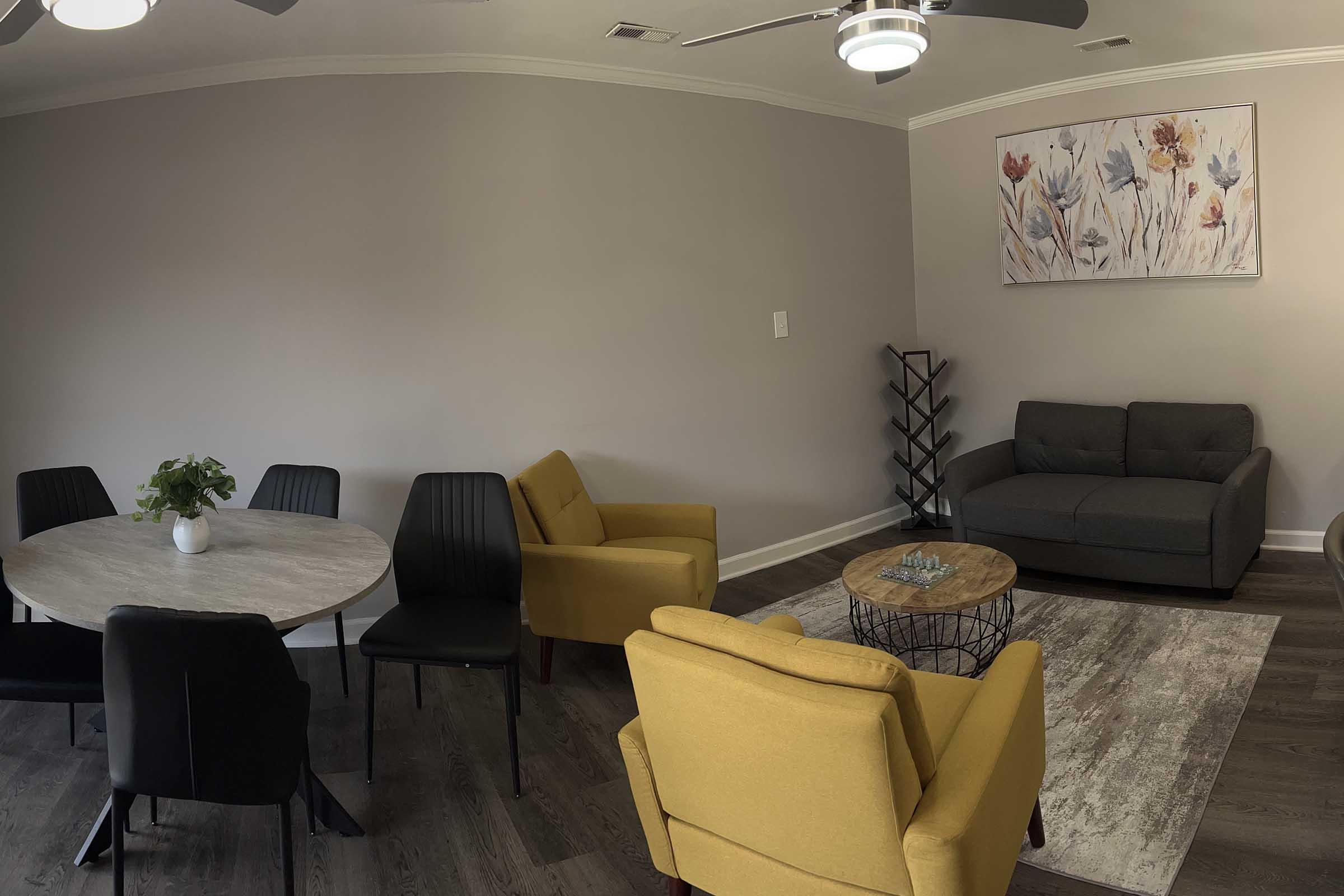 A cozy living area featuring a gray sofa, two yellow armchairs, and a round dining table with black chairs. A potted plant sits on the table. There is a multi-tiered shelf against the wall and a light-colored rug on the floor. A large artwork with floral designs hangs above the sofa, and a ceiling fan is visible.