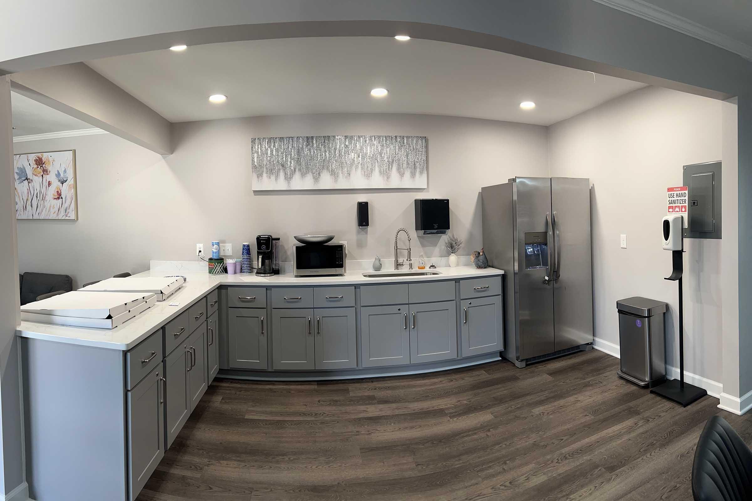 A modern kitchen featuring gray cabinetry, a white countertop, a stainless steel refrigerator, a microwave, and a sink. The space includes a coffee station and a soap dispenser. The walls are light-colored, decorated with a floral artwork, and the floor is dark wood.