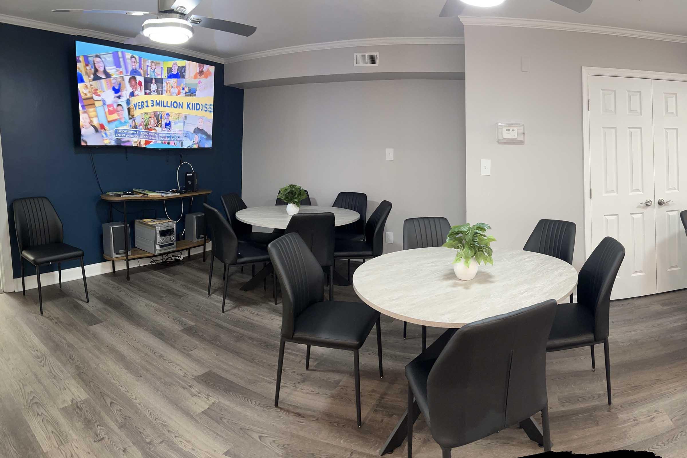 A modern meeting room featuring several round tables surrounded by black chairs. A flat-screen TV is mounted on the wall, displaying colorful images and text related to kids' education. The room has a sleek, minimalist design with light-colored walls and a wooden floor, complemented by small green plants on the tables.