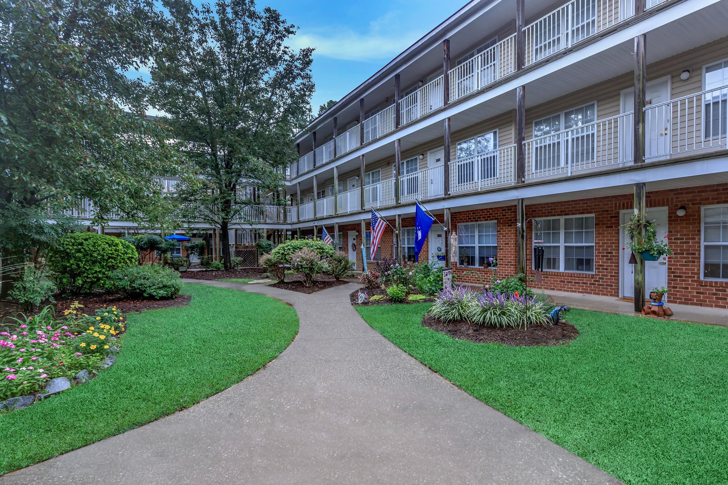 A well-maintained courtyard of a multi-story building, featuring landscaped gardens with colorful flowers, a pathway winding through the greenery, and flags displayed near the entrance. The architecture includes balconies, and the scene is set under a clear blue sky.