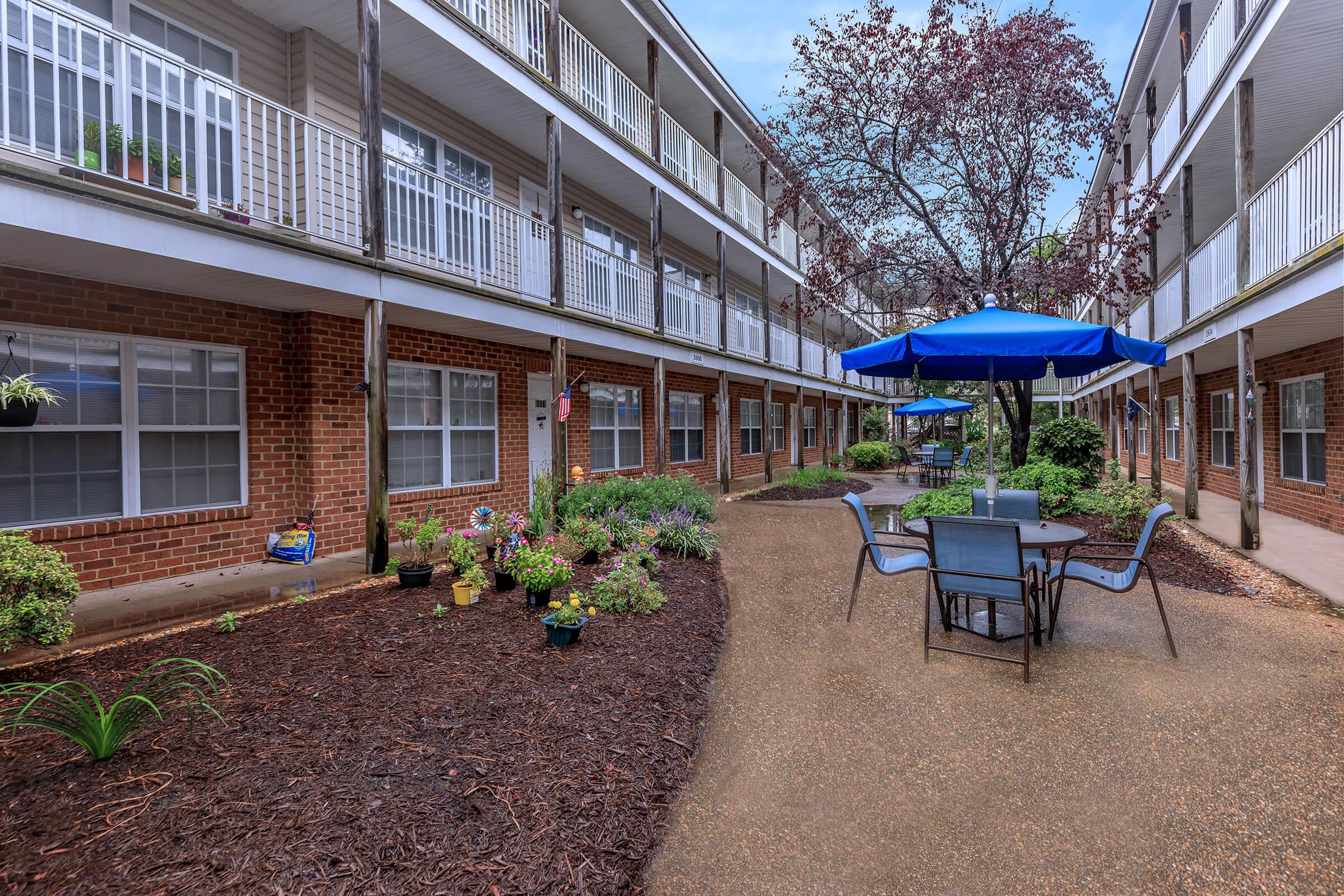A view of a courtyard area between two multi-story brick apartment buildings. The space features a round table with chairs beneath a blue umbrella, surrounded by landscaped gardens with various plants and flowers. The scene is peaceful and inviting, with a clear sky above.