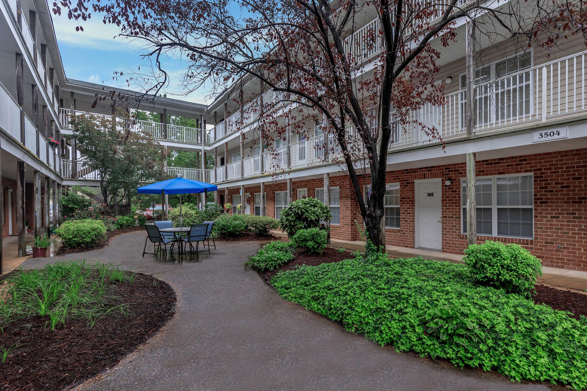 A peaceful courtyard in an apartment complex featuring a blue umbrella over a round table with chairs. Lush greenery surrounds the pathway, and there are several small shrubs and trees, creating a serene outdoor space. The building has multiple balconies, contributing to the community atmosphere.