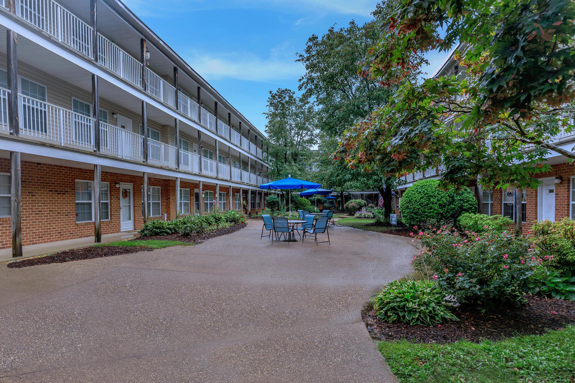 A tranquil courtyard featuring a round table and chairs under a blue umbrella, surrounded by well-maintained greenery and flower beds. The scene includes two levels of brick buildings with balconies, creating a peaceful outdoor space for relaxation.
