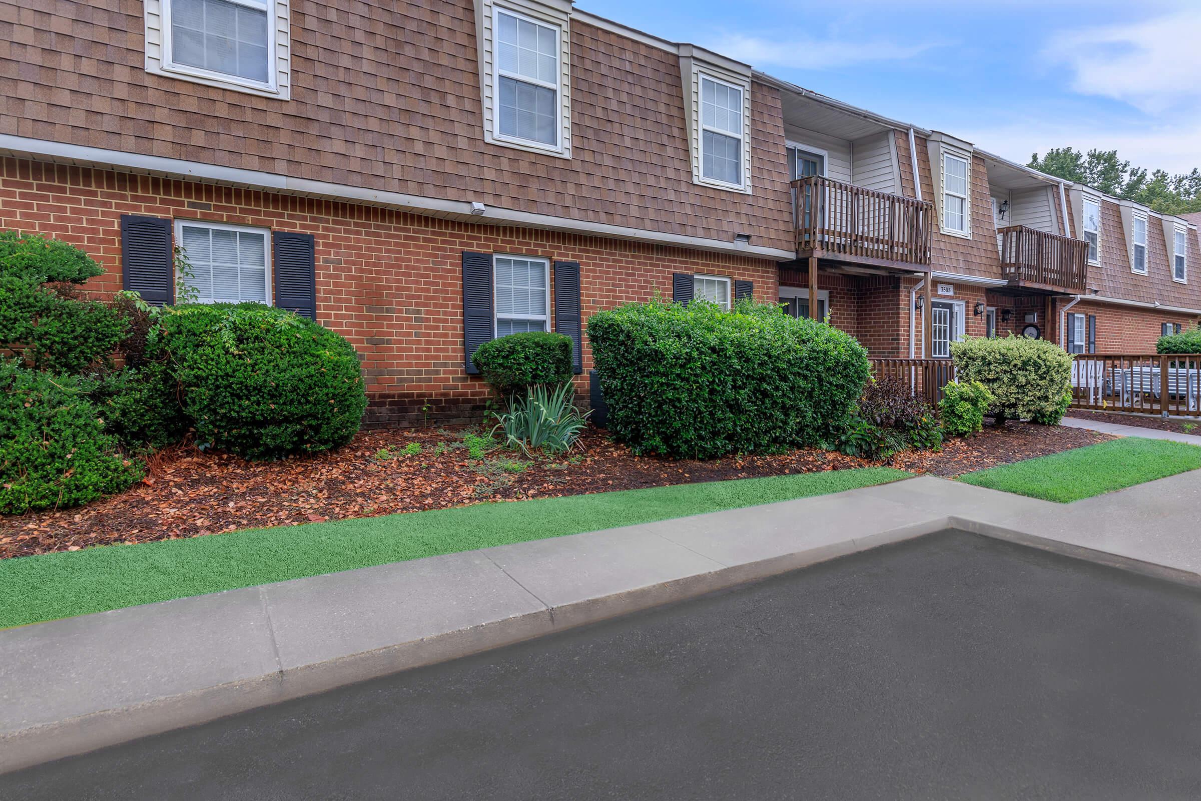 A view of a residential building with brick exterior featuring multiple units. The area has neatly trimmed bushes and green lawn in the foreground, along with a sidewalk leading to the entrance. Balconies are visible above, and the sky is clear in the background.