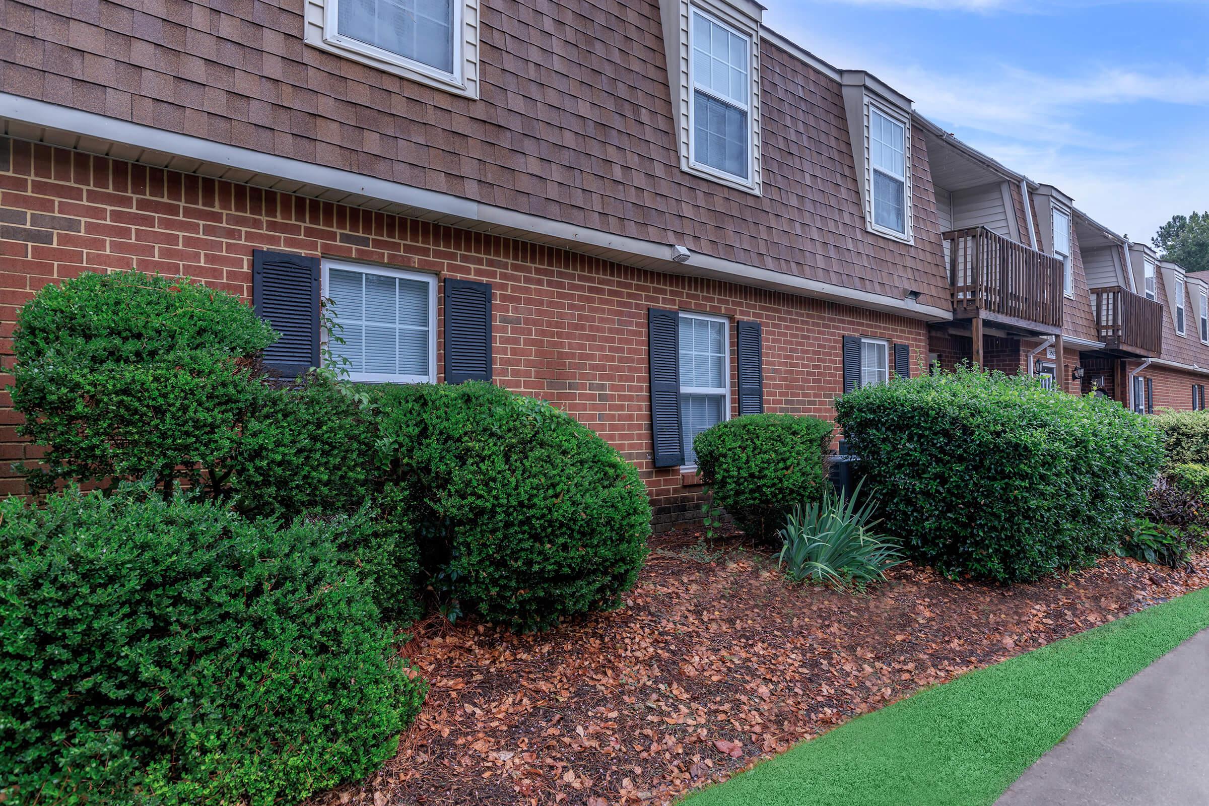 A landscaped area in front of a brick apartment building featuring neatly trimmed bushes, small plants, and brown mulch along a paved walkway. The building has multiple windows framed by dark shutters and a textured brown shingle roof, with a clear blue sky above.