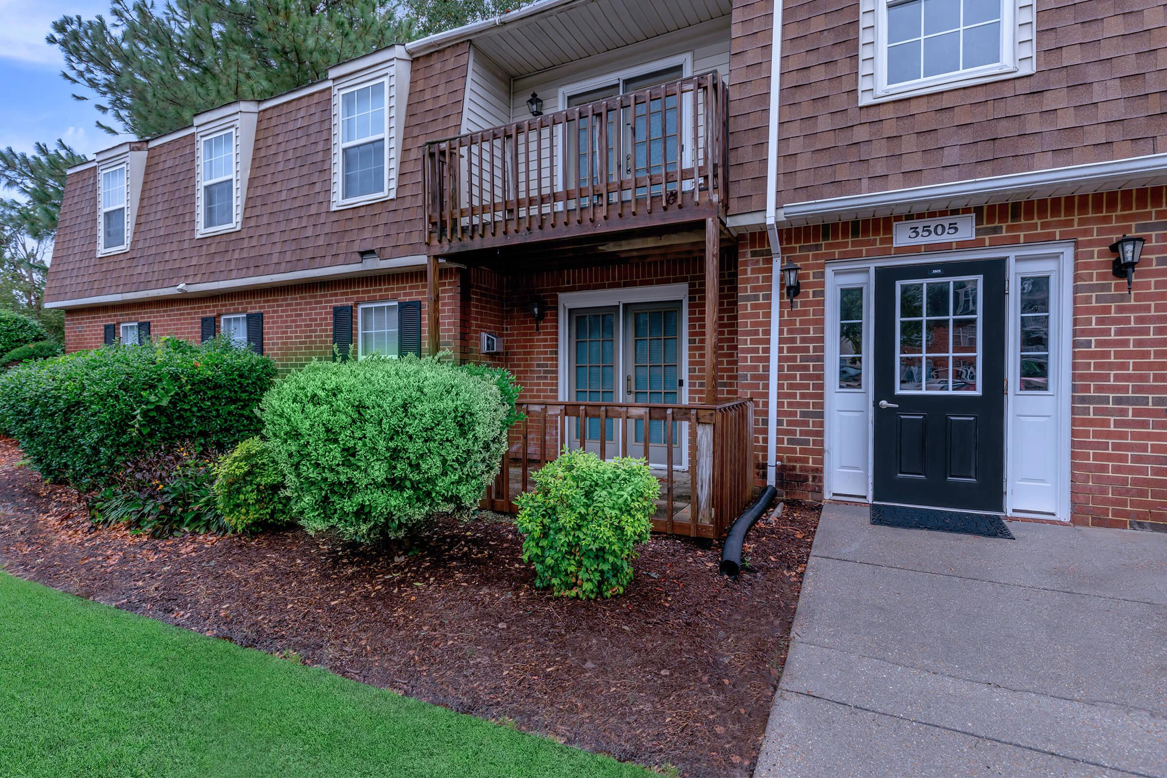 A two-story brick apartment building with a small wooden balcony on the second floor, surrounded by neatly trimmed bushes and green grass. The entrance features a black door with glass panes, and there are windows above it. The setting is outdoor with well-maintained landscaping.