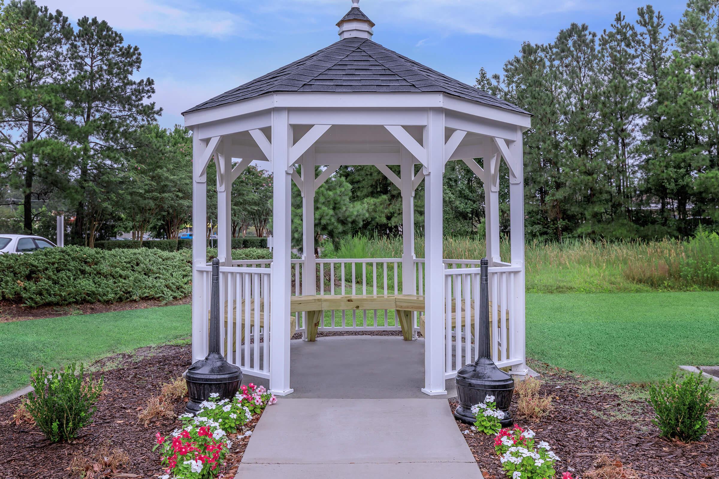 A white gazebo with a black roof, surrounded by green grass and landscaped flower beds. The structure features an open design with a seating area inside. In the background, trees and shrubs create a natural setting.
