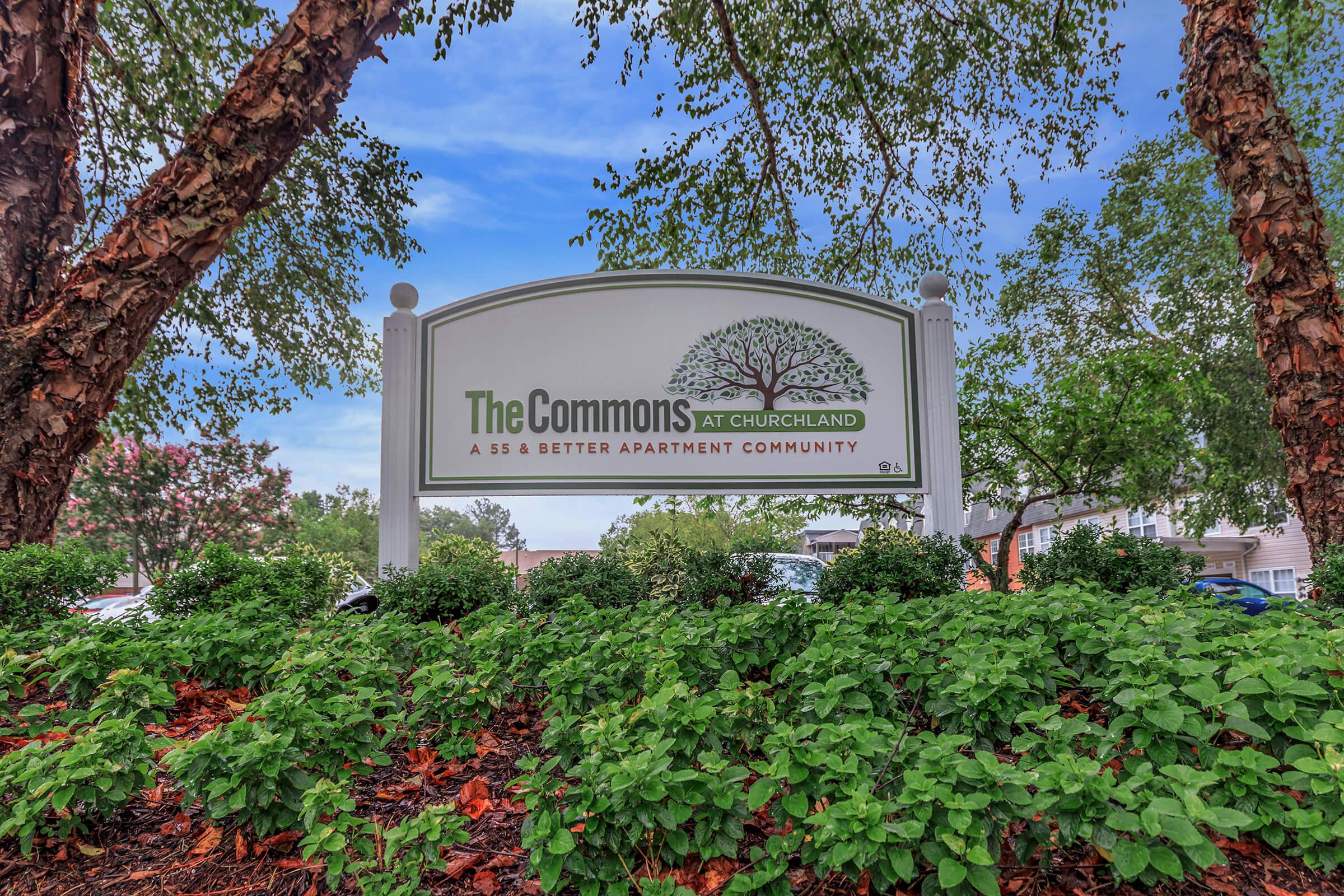 Sign for "The Commons at Churchland," indicating it's a 55 and better apartment community. Surrounded by greenery, including shrubs and trees, with a blue sky in the background.