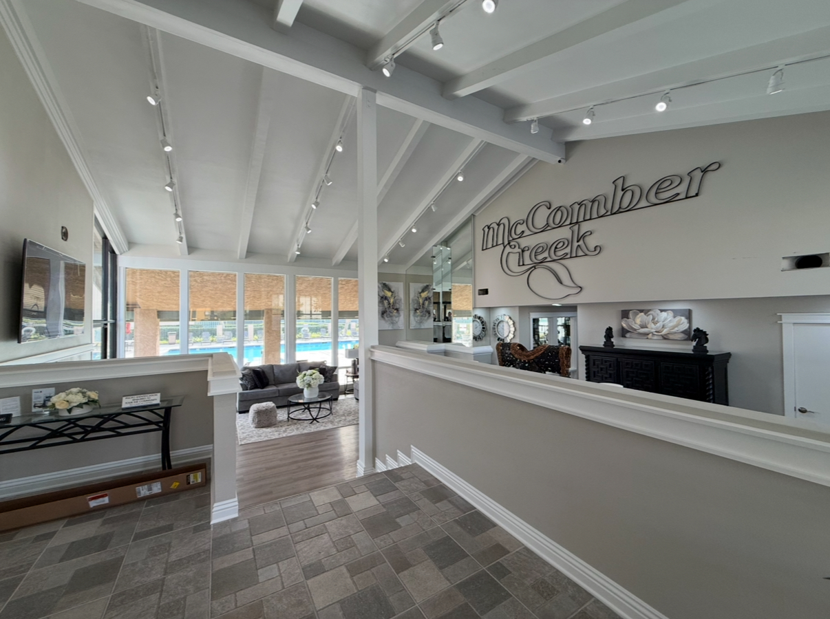 A modern entrance area with high, beamed ceilings and stylish decor. The walls feature the name "McComber Creek" in an elegant font. Large windows allow natural light to flood the space, revealing a view of a pool outside. The room is furnished with comfortable seating and decorative accents, creating a welcoming atmosphere.