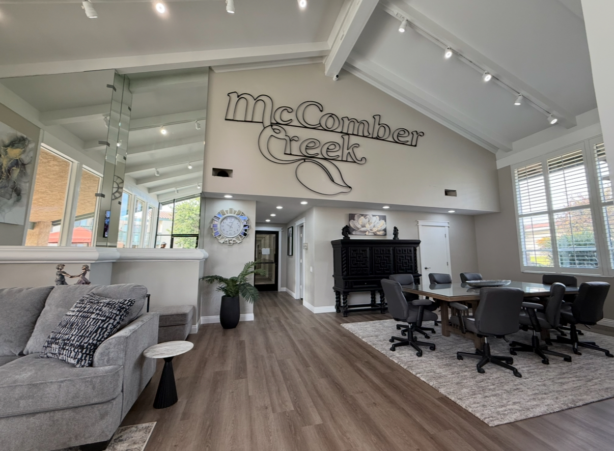 Interior view of a modern living space featuring a spacious layout with a gray sofa, a large wooden dining table surrounded by black chairs, and a decorative wall displaying the name "McComber Creek." The space has high ceilings, large windows allowing natural light, and contemporary decor elements.