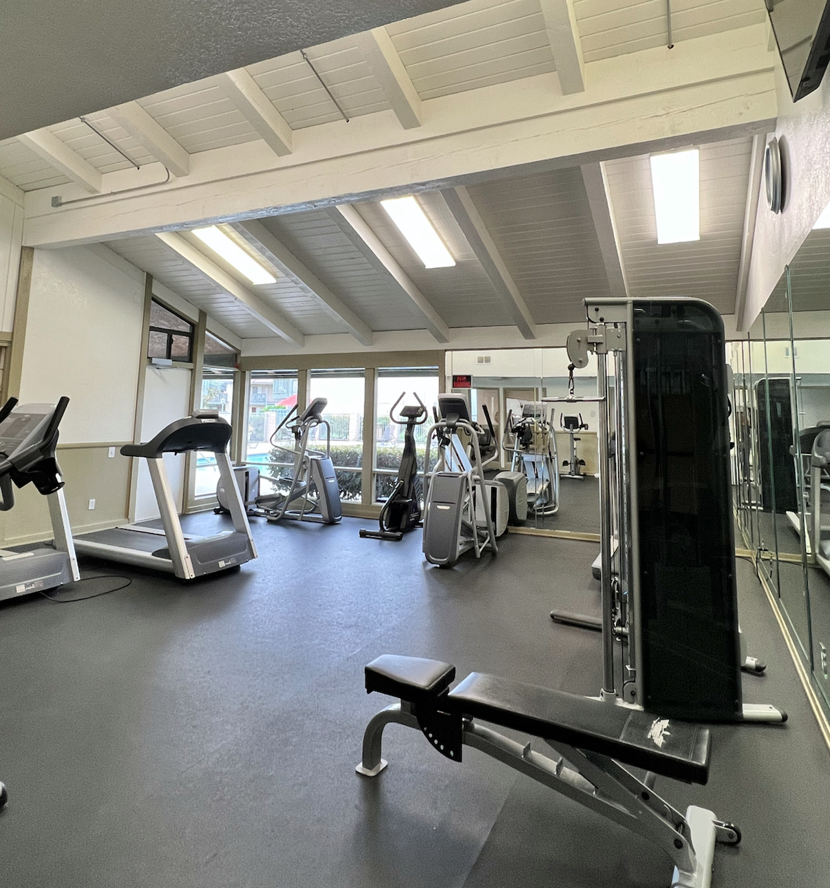 Interior of a gym featuring various exercise equipment, including treadmills, stationary bikes, and weight benches. The space has high ceilings with beams, ample lighting, and mirrors on one wall, creating a bright and open atmosphere for workouts.