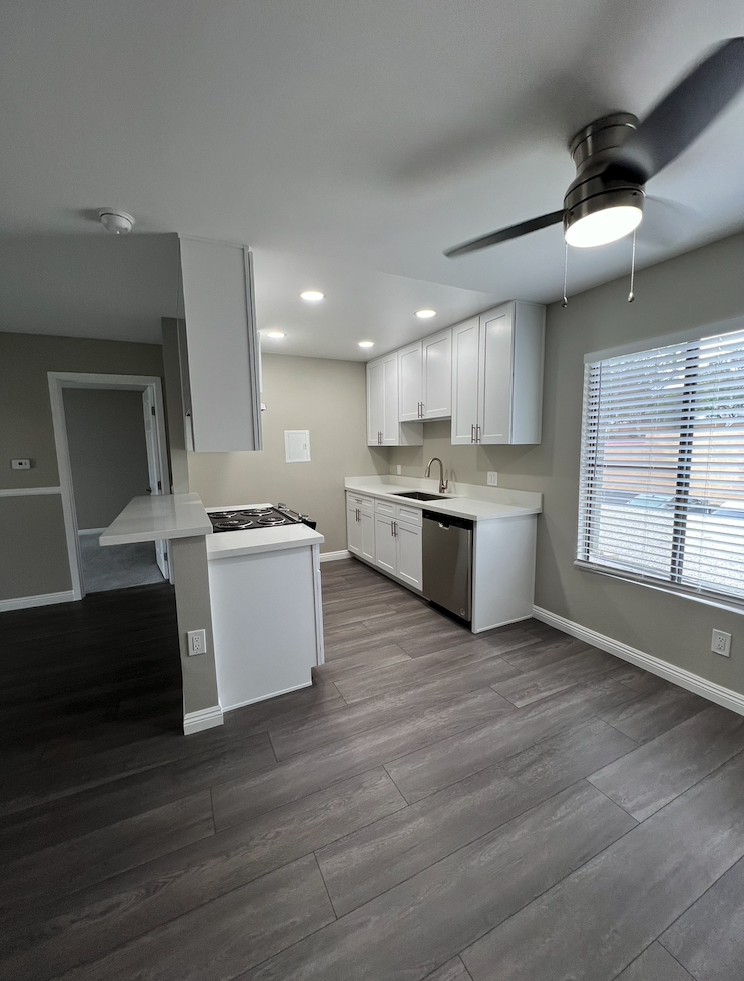 Modern kitchen featuring sleek white cabinets, a stainless steel dishwasher, and a gas stove. The space has a light gray wall color and wood-look tile flooring. A ceiling fan provides ventilation, and there is a window with blinds allowing natural light into the room.