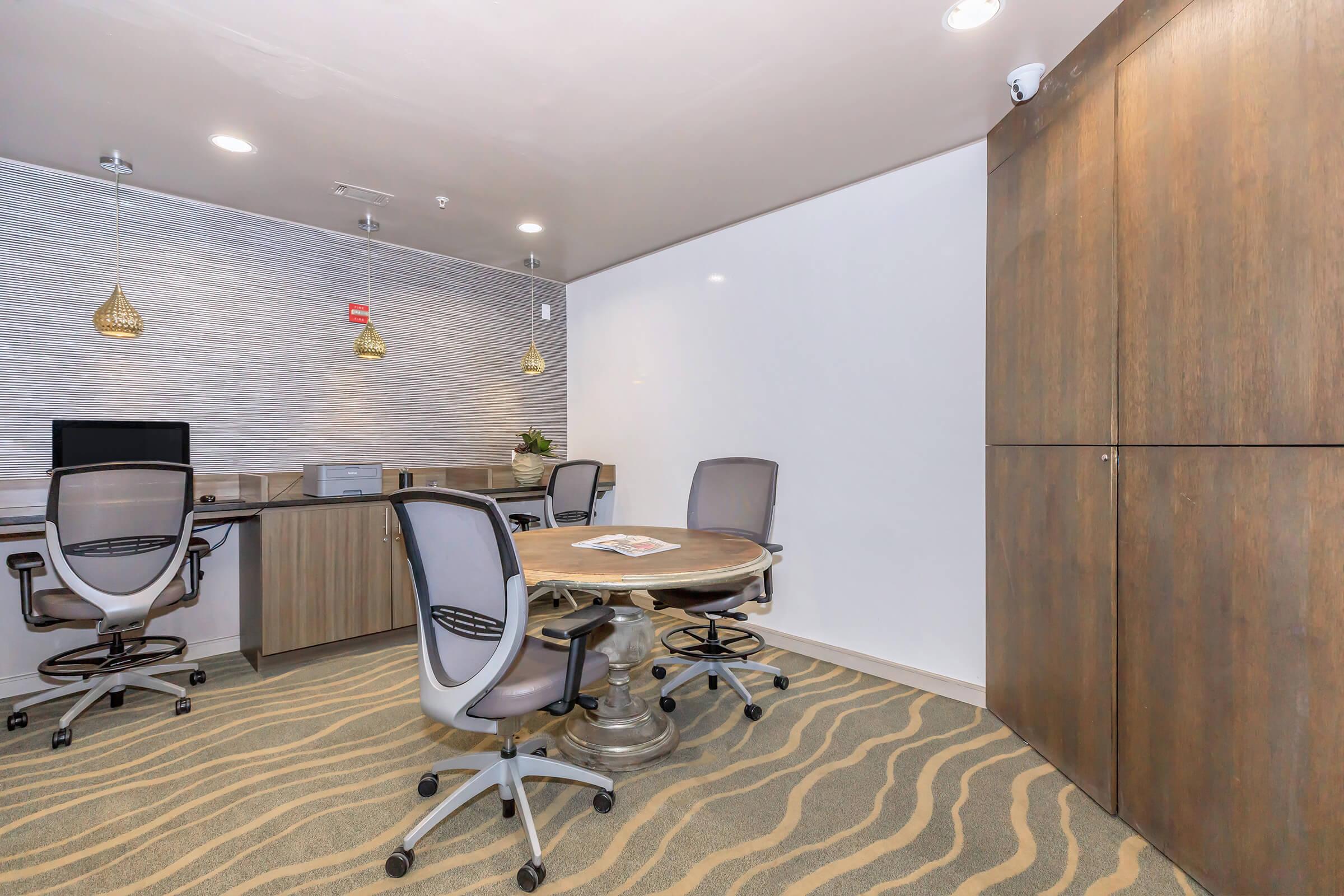 A modern office interior featuring a round table with four chairs, two computer workstations, and a wooden cupboard. The walls have a textured design, and stylish pendant lights hang from the ceiling. The carpet has a wavy pattern, adding to the contemporary aesthetic of the space.