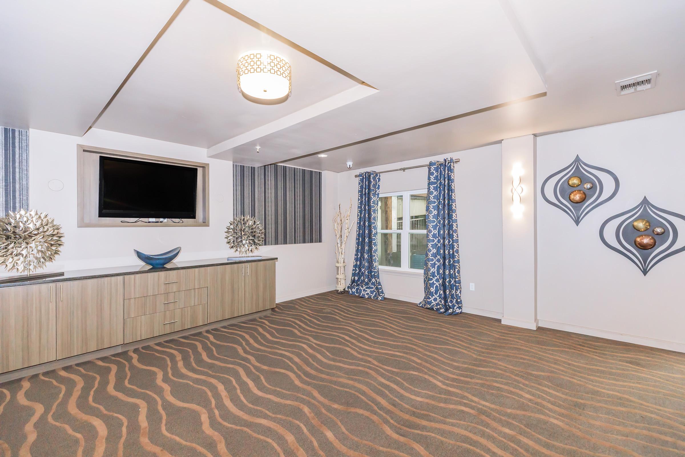 A modern interior space featuring a stylish design. The room includes a television mounted on the wall, a dresser with decorative objects, and patterned curtains. The carpet has a wavy design, and there are decorative wall accents with circular elements in gold tones. Natural light enters through a window.