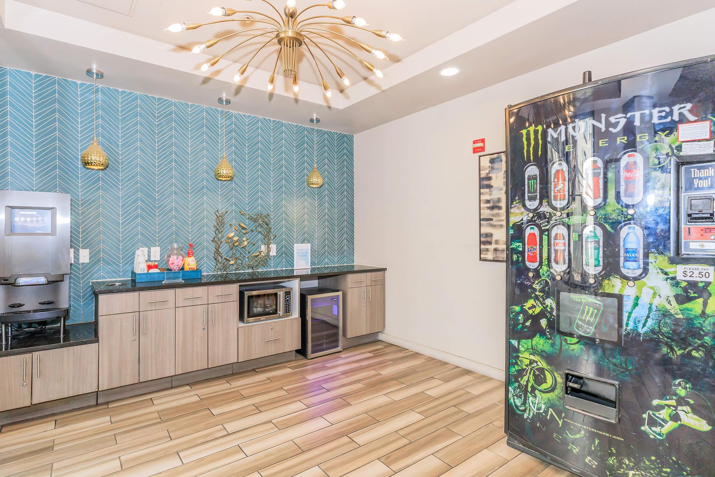 A modern break room featuring a patterned blue wall, a countertop with a microwave and refrigerator, and a vending machine stocked with energy drinks. Decorative lights hang from the ceiling, contributing to a contemporary atmosphere.
