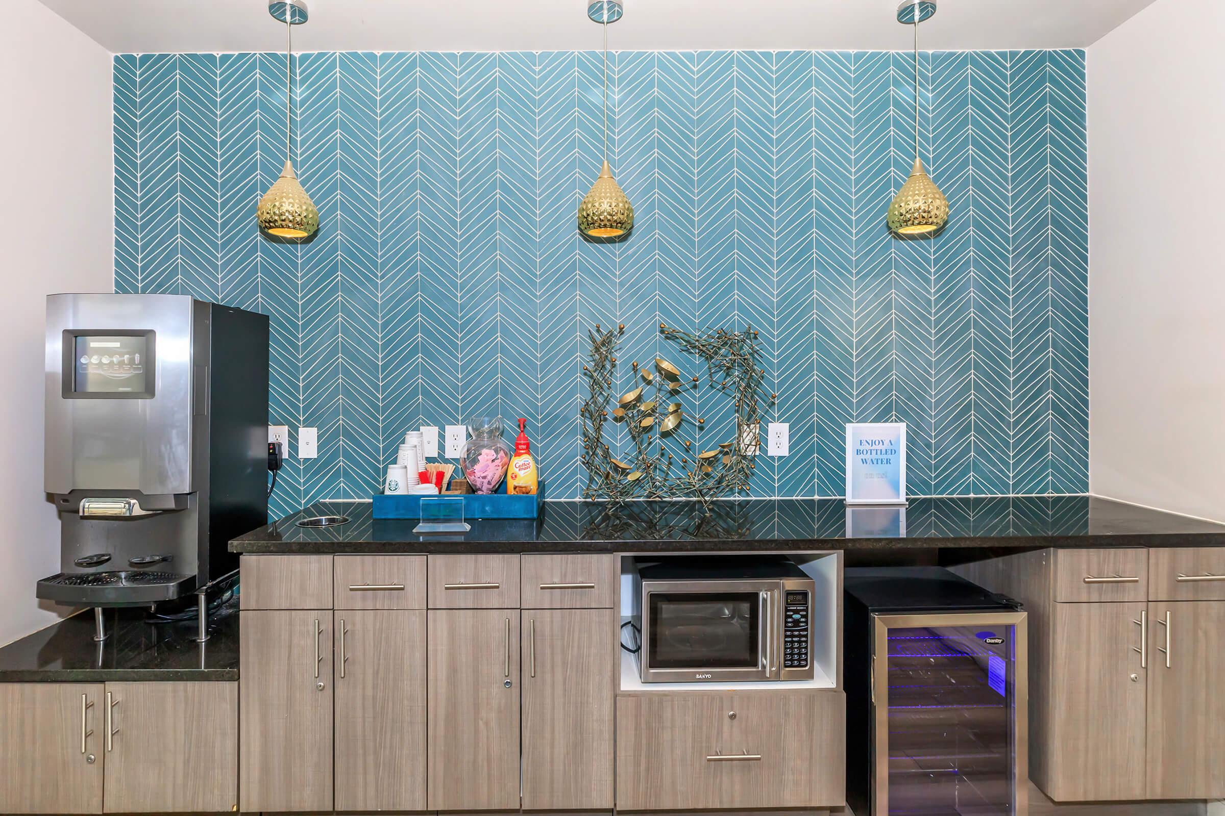 Modern kitchen area featuring a teal herringbone-patterned wall, pendant lighting, a coffee machine, and a microwave. A countertop holds various condiments and a mini fridge, while a decorative arrangement made of branches is displayed. A sign reads "ENJOY YOUR WATER."