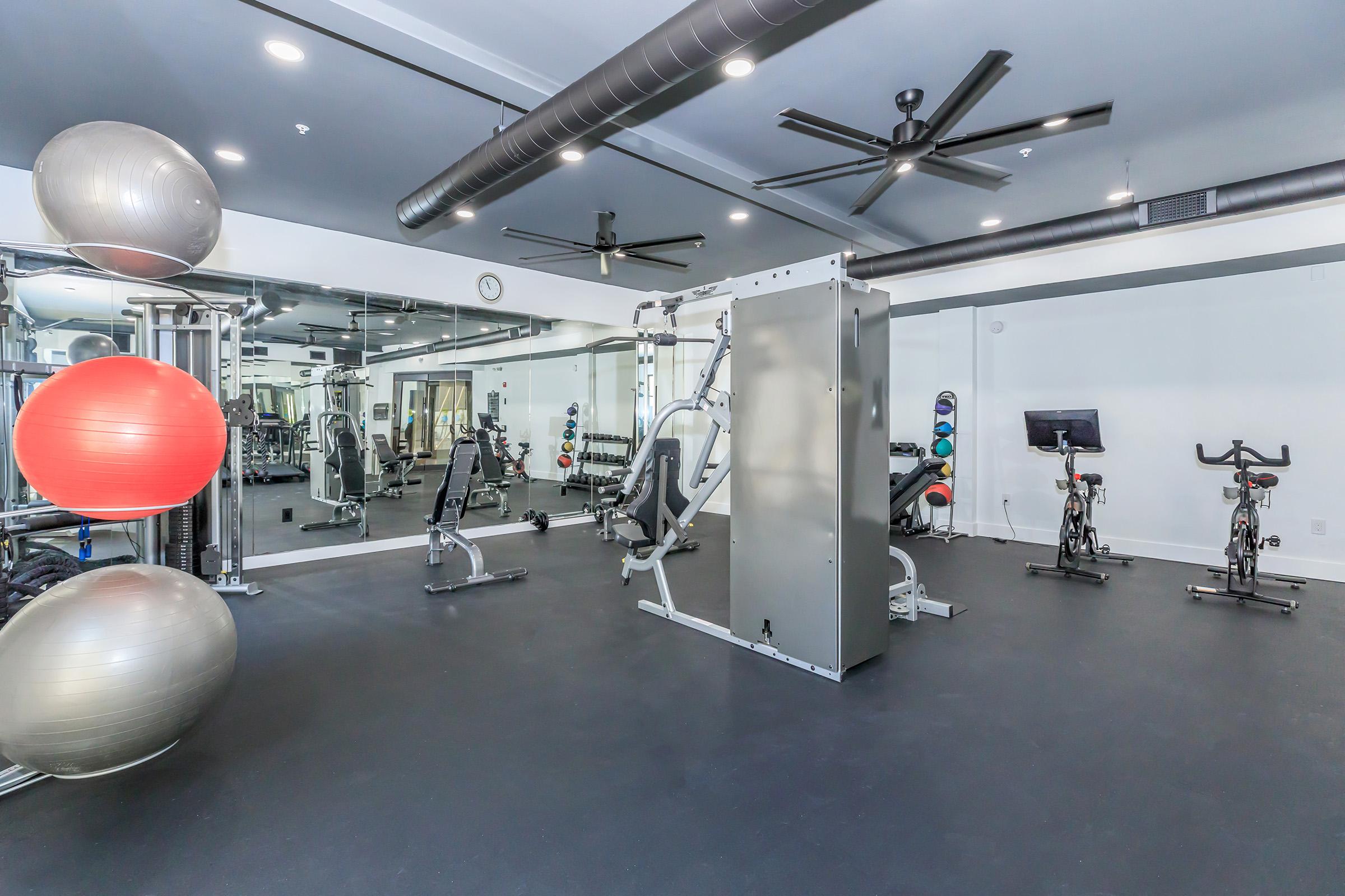 A modern gym space featuring exercise equipment, including weight machines, stationary bikes, and large exercise balls. The room has a mirror on one wall, ceiling fans, and is well-lit, creating an inviting atmosphere for workouts.