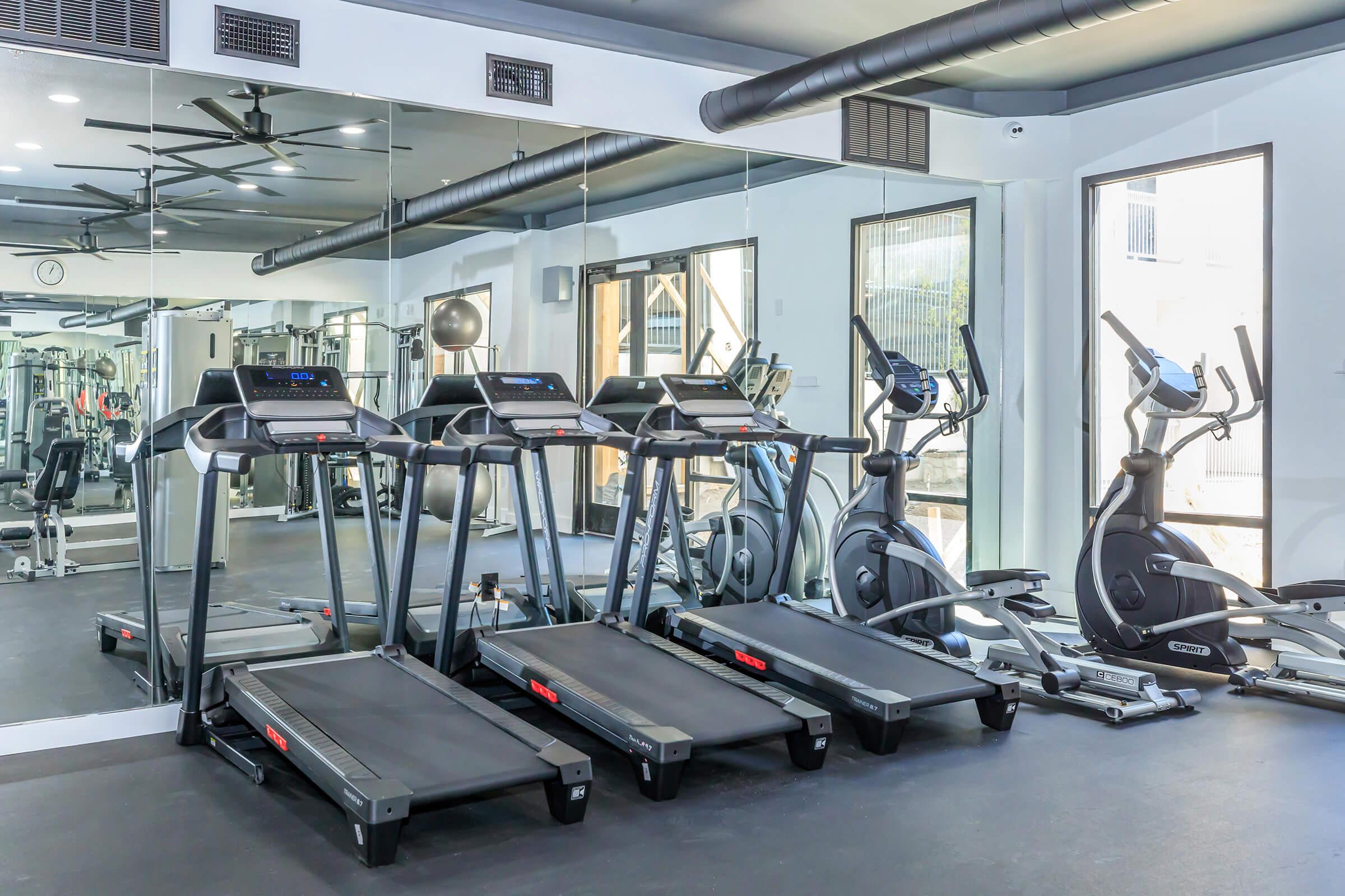 A modern gym interior featuring three treadmills and an elliptical machine. The space is well-lit with large mirrors on the wall, and various gym equipment can be seen in the background. The flooring is dark, contributing to a sleek and clean atmosphere.