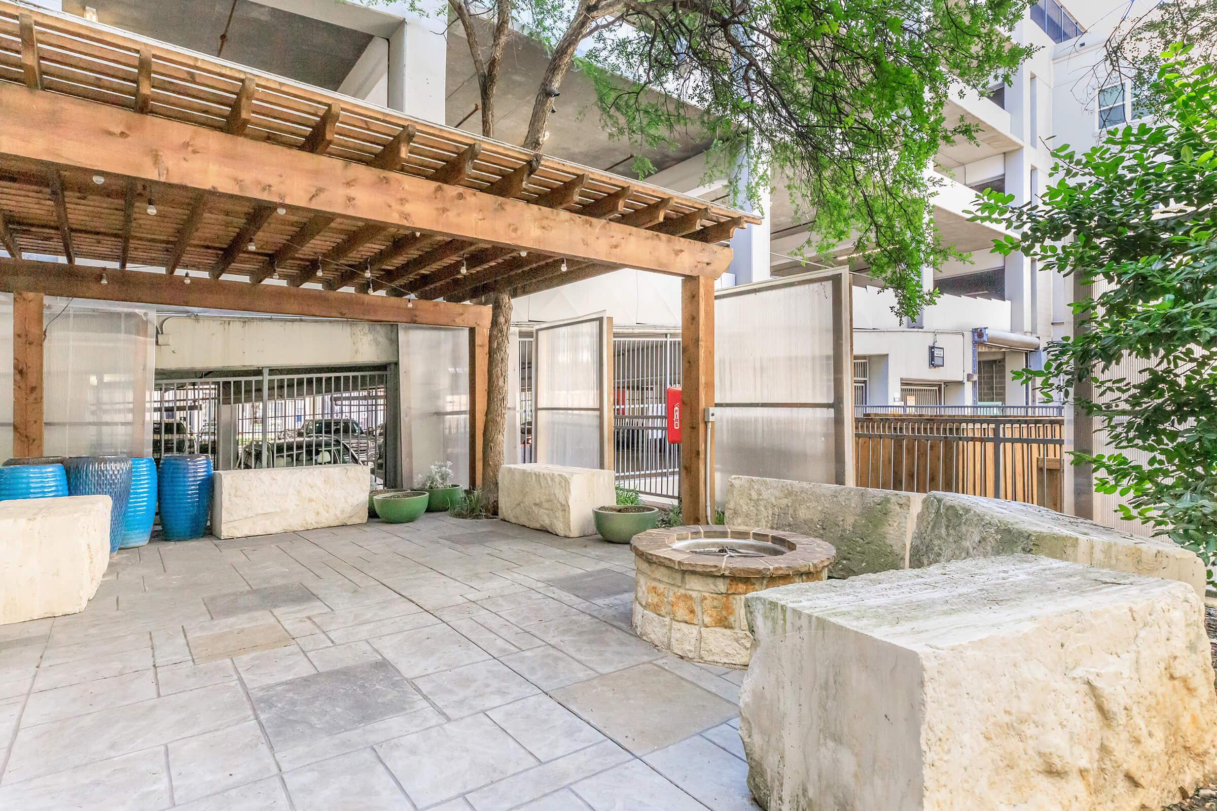 Outdoor patio area with a wooden pergola, stone seating, and a circular fire pit. The space is surrounded by greenery and has a gated entrance. Potted plants are visible, and part of a parking structure can be seen in the background.
