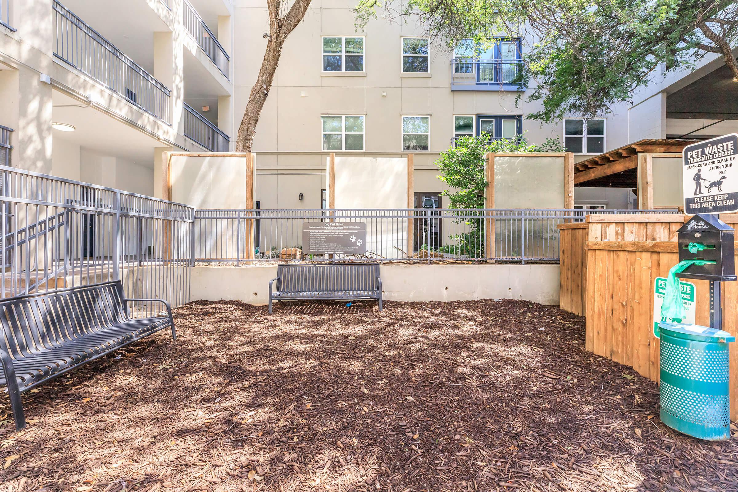 A pet-friendly area featuring benches, mulch ground cover, and waste disposal stations. The space is partially enclosed with wooden accents and greenery in the background, set in a residential building environment.