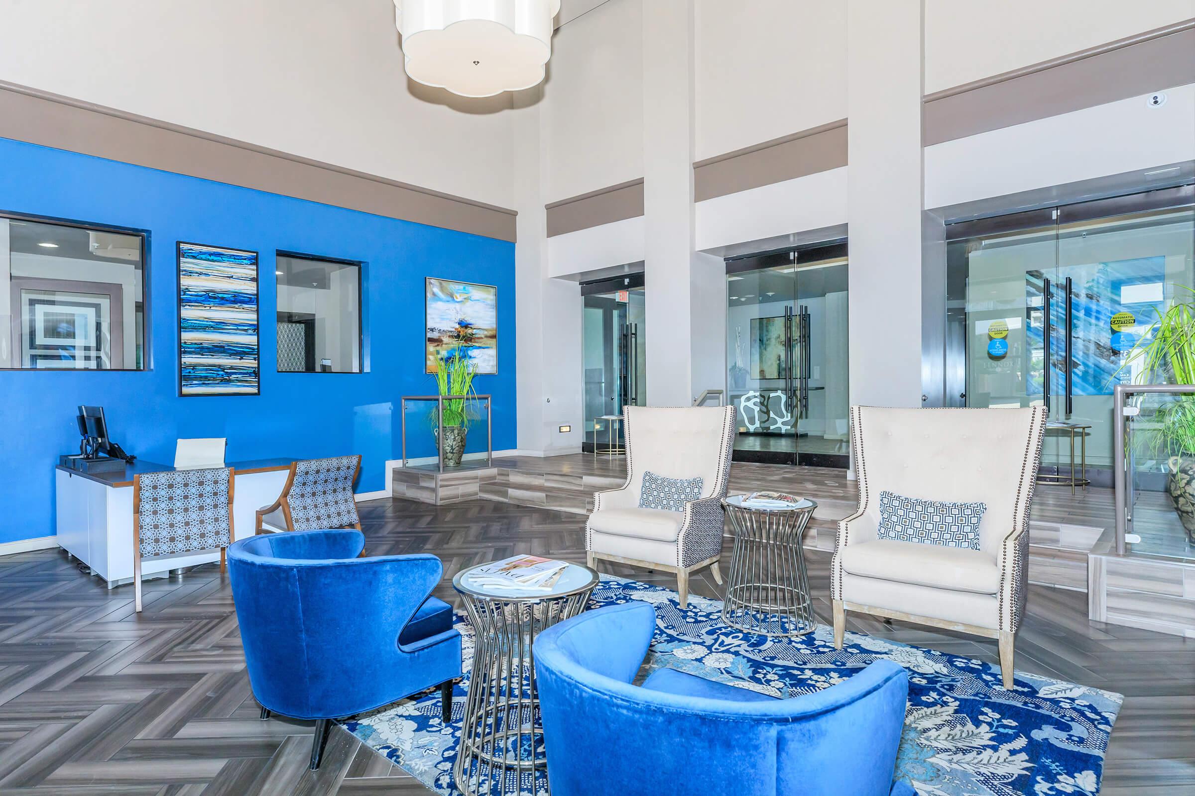 Modern lobby with blue accent walls, featuring comfortable chairs and a stylish rug. There are large windows allowing natural light, and artistic wall decor. A reception desk is positioned in the background. The space is clean and inviting, designed for relaxation and social interaction.