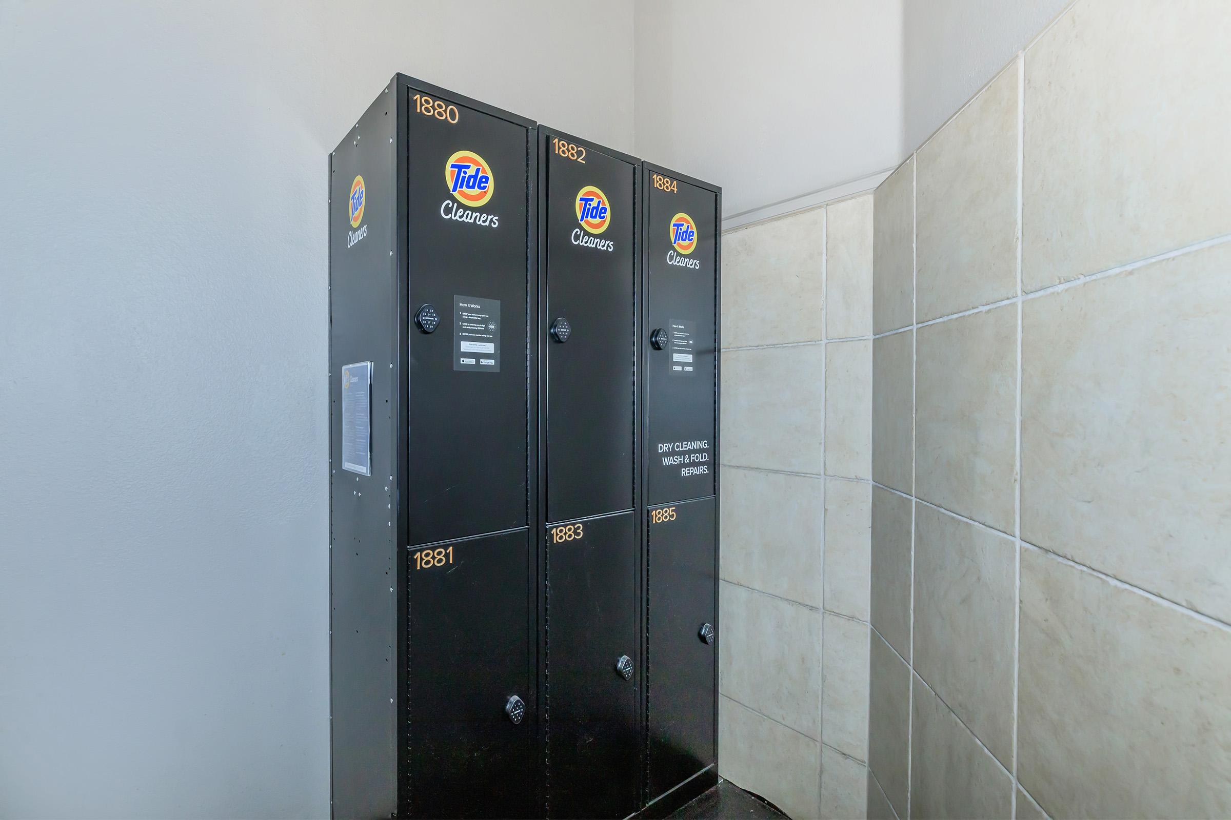 A row of black lockers branded with Tide Cleaners logos in a corner of a tiled room. Each locker is labeled with a number, and a directory or instruction sheet is visible on the left side. The environment appears well-lit and clean, suggesting a laundry or dry-cleaning facility.