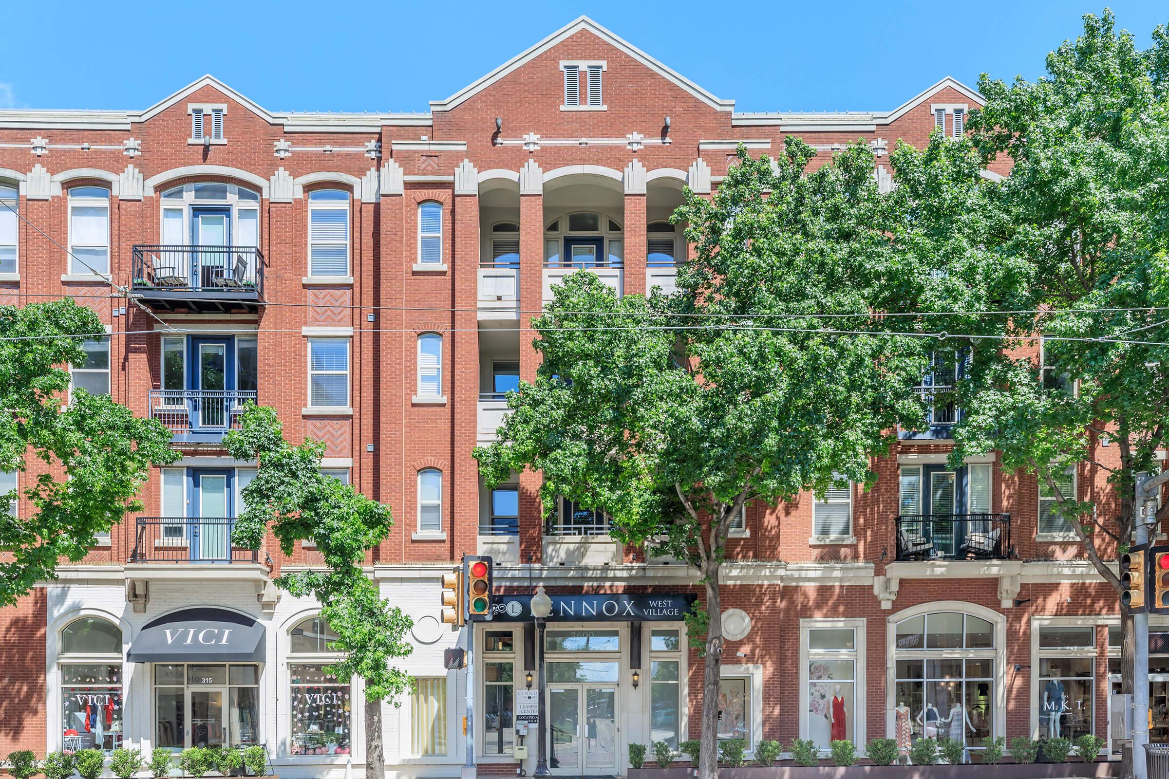 A red brick apartment building featuring multiple balconies, large windows, and decorative arches. Below, there are storefronts with signs. Lush green trees line the sidewalk, and a traffic light is visible in the foreground under a clear blue sky.