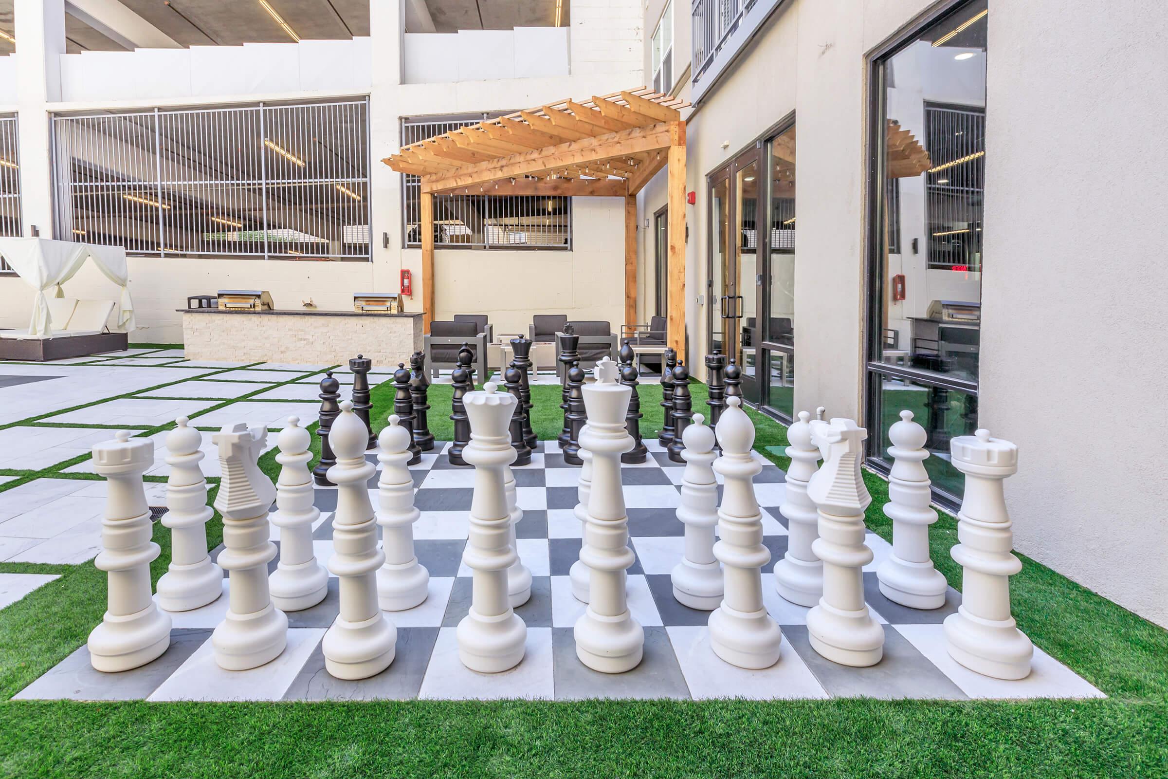 A large outdoor chess set with oversized black and white pieces arranged on a checkerboard pattern. The setting features a modern building in the background, with green grass and shaded seating areas nearby. The scene is bright and inviting, perfect for playing chess outdoors.