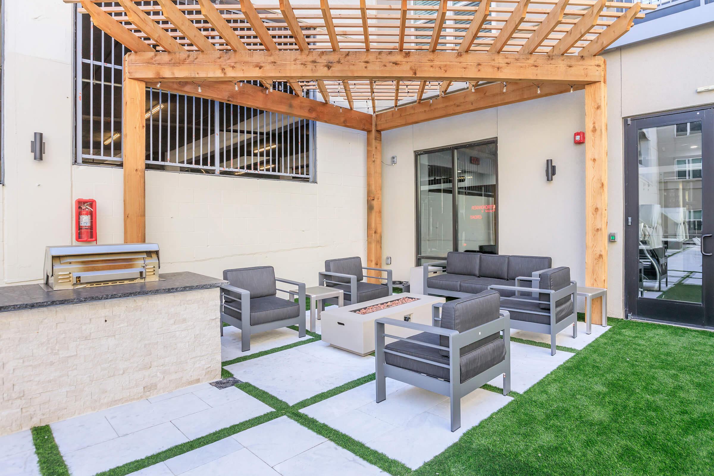 An outdoor lounge area featuring gray upholstered seating arranged around a fire pit. There's a decorative wooden pergola overhead, and the flooring has a mix of tiles and artificial grass. A barbecue grill is situated on one side, complementing the modern and inviting ambiance.