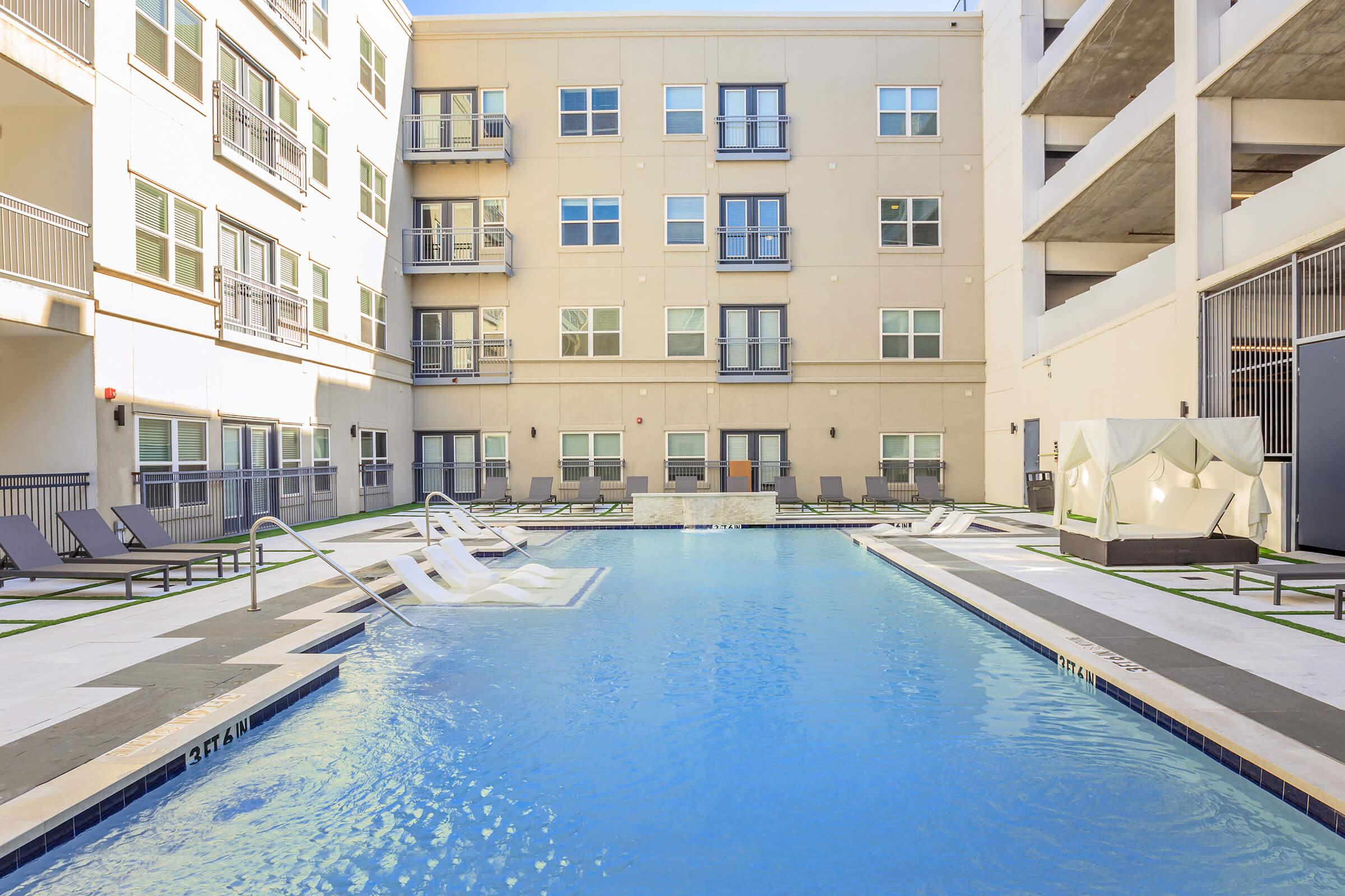 A serene outdoor swimming pool surrounded by lounge chairs, situated in a modern apartment complex courtyard. The pool features a water fountain and is bordered by clean, minimalist architecture, with multiple balconies visible in the background. Sunlight reflects off the water, creating a relaxing atmosphere.