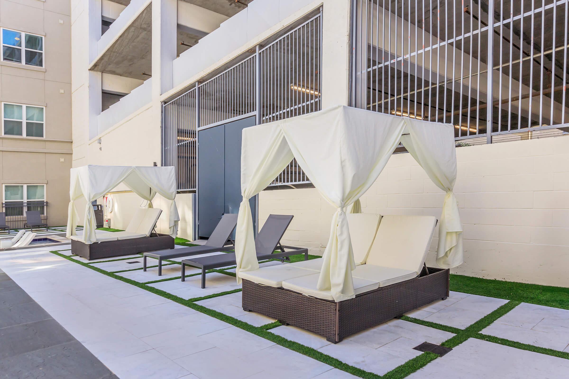 Two white canopied lounge beds are placed on a stone patio with artificial grass, surrounded by a modern building and a parking garage. One lounge chair is gray and the other is empty. The area appears inviting and well-maintained, ideal for relaxation.