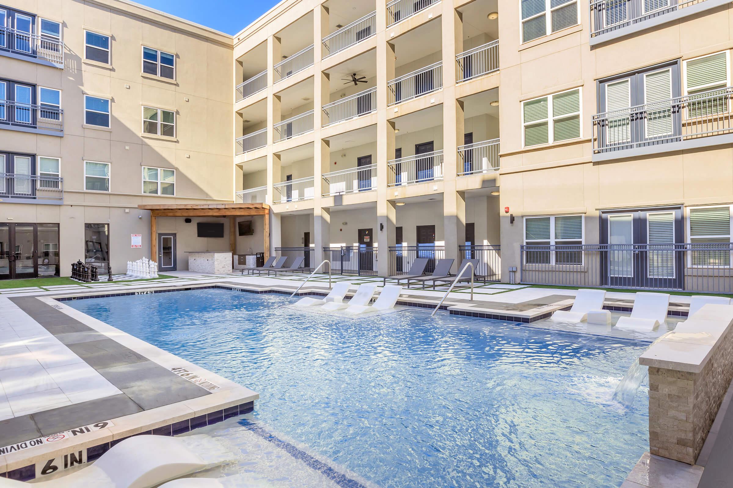 A modern swimming pool surrounded by lounge chairs, with a multi-story apartment building in the background. The pool area features a hot tub and is designed with stone and tile accents, under a clear blue sky.