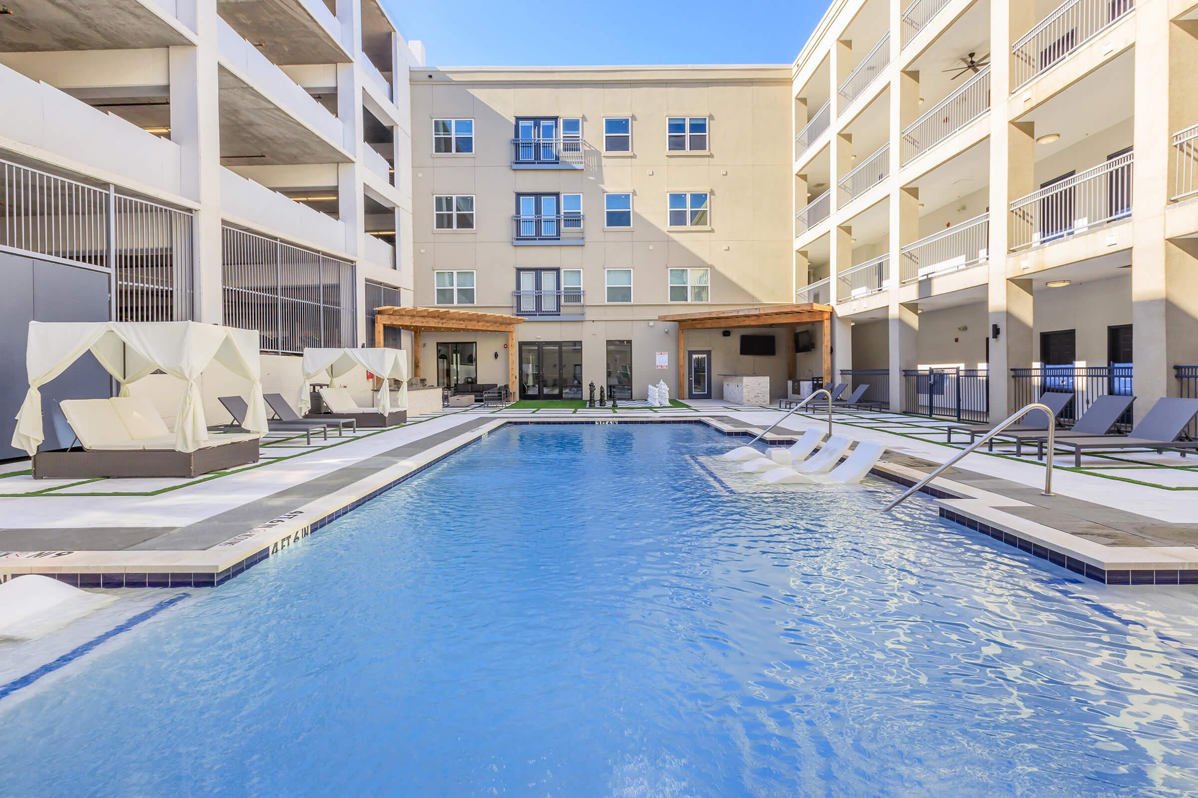 A modern outdoor pool area with lounge chairs and cabanas, surrounded by a sleek residential building. The pool features a gentle slope leading into the water, and the space is well-lit with natural sunlight, offering a relaxing atmosphere for residents.