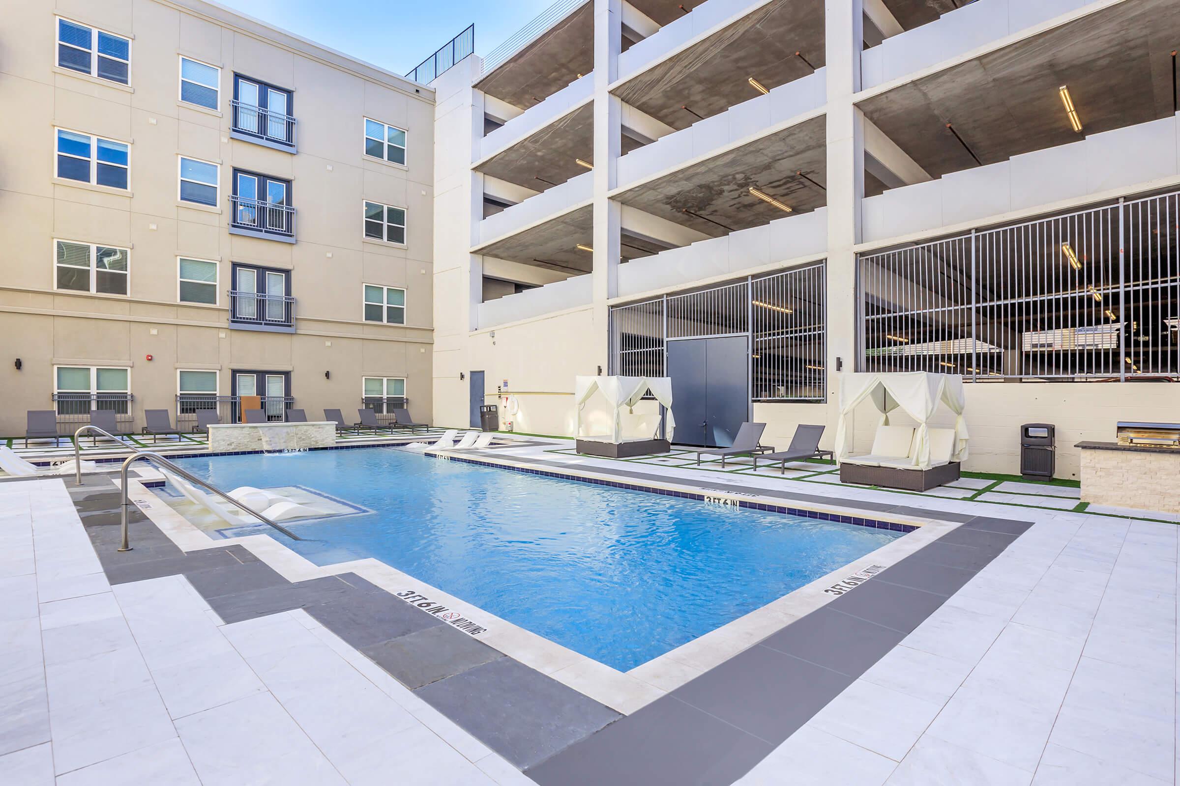 A modern apartment complex featuring a swimming pool surrounded by lounge chairs and cabanas. The pool area is located next to a multi-level parking garage, with clean white tiles and a landscaped space for relaxation. Windows of nearby apartments overlook the pool area.