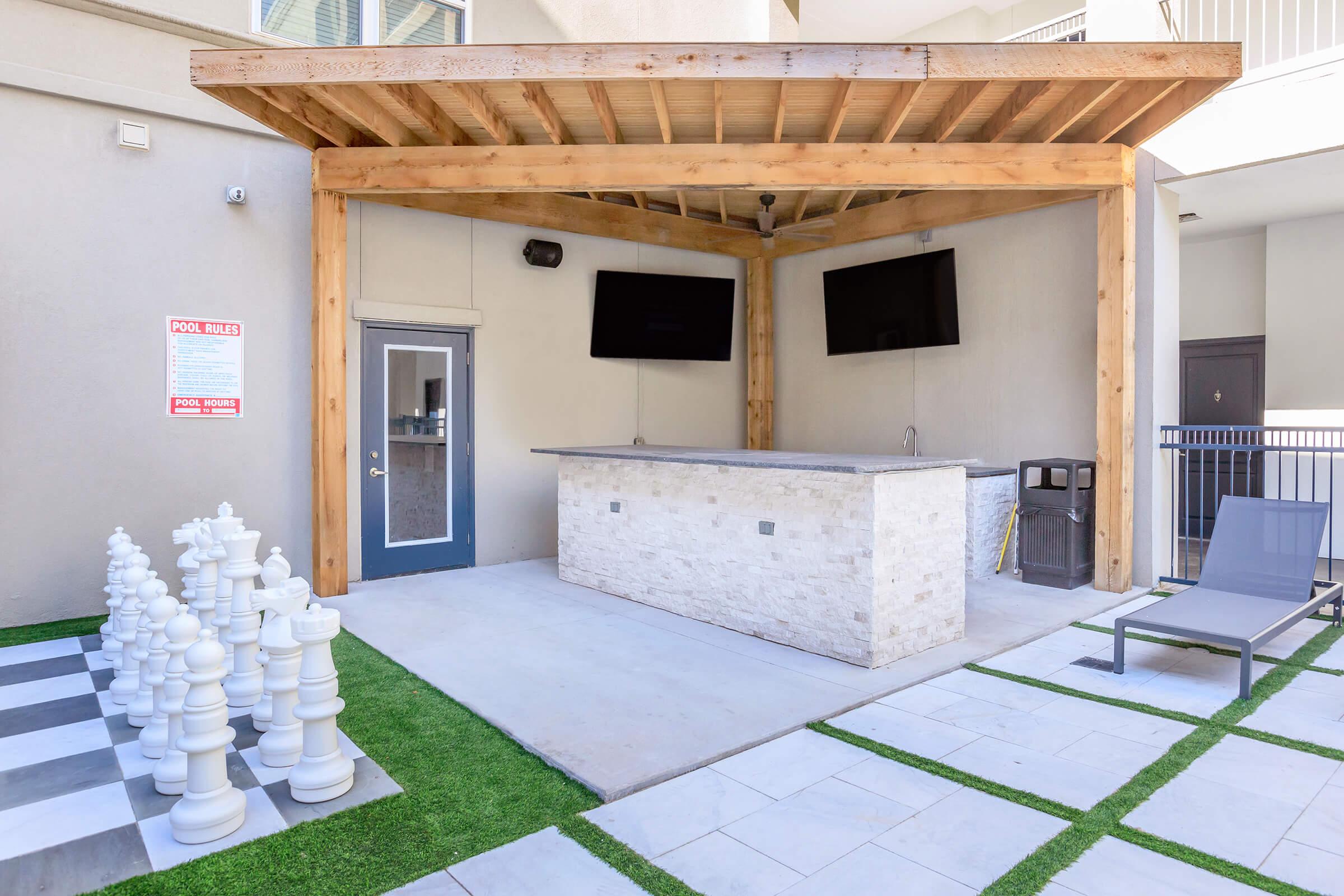 An outdoor space featuring a covered bar area with two mounted TVs, a trash bin, and a lounge chair. In front, there is a large chessboard with white chess pieces on a green lawn surface, creating a relaxing atmosphere for leisure and entertainment.