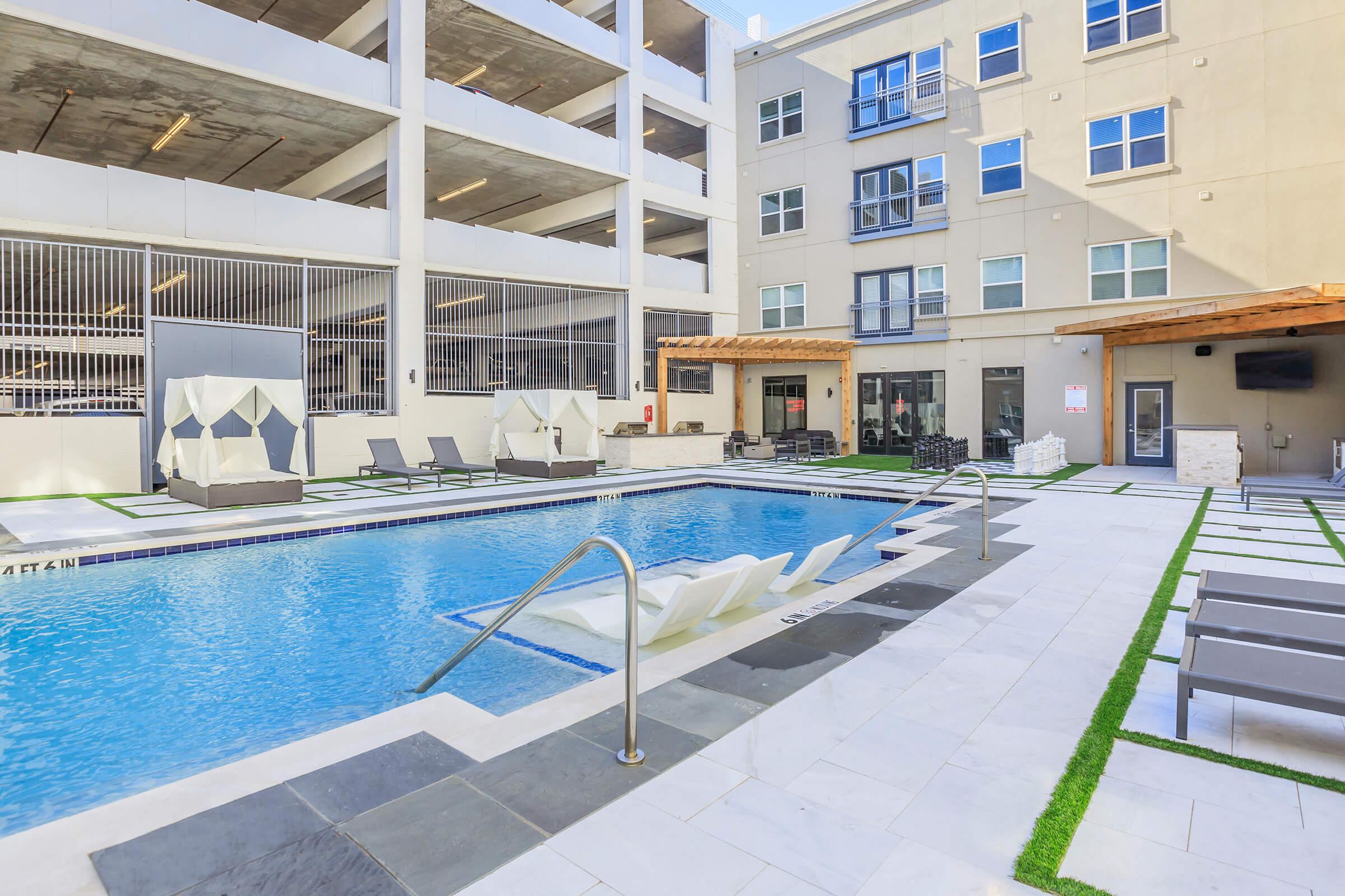 A modern outdoor pool area featuring lounge chairs and umbrellas, surrounded by green grass. The backdrop includes a multi-level parking structure and an apartment building with large windows. The pool area is designed for relaxation and leisure, creating a welcoming environment.
