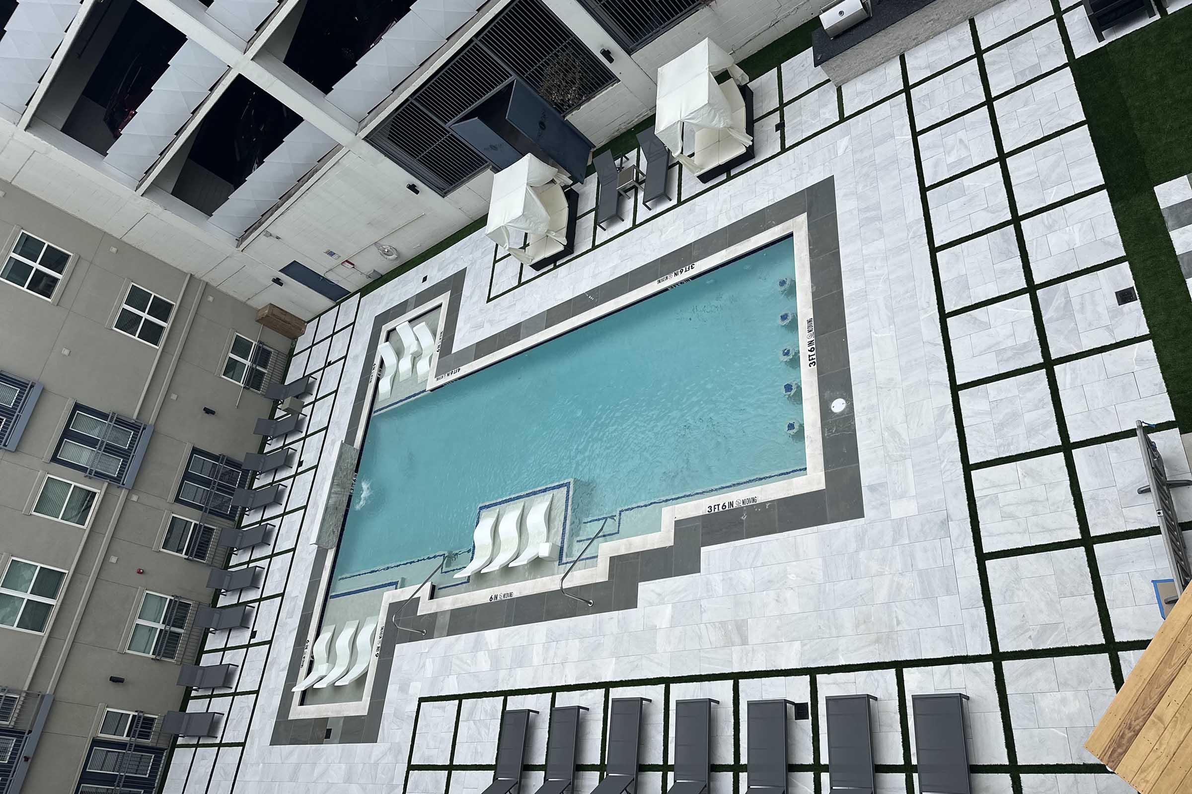 Aerial view of a modern swimming pool surrounded by lounge chairs and cabanas, featuring a sleek, geometric design. The pool area is bordered by light-colored tiles and greenery, with buildings visible in the background.