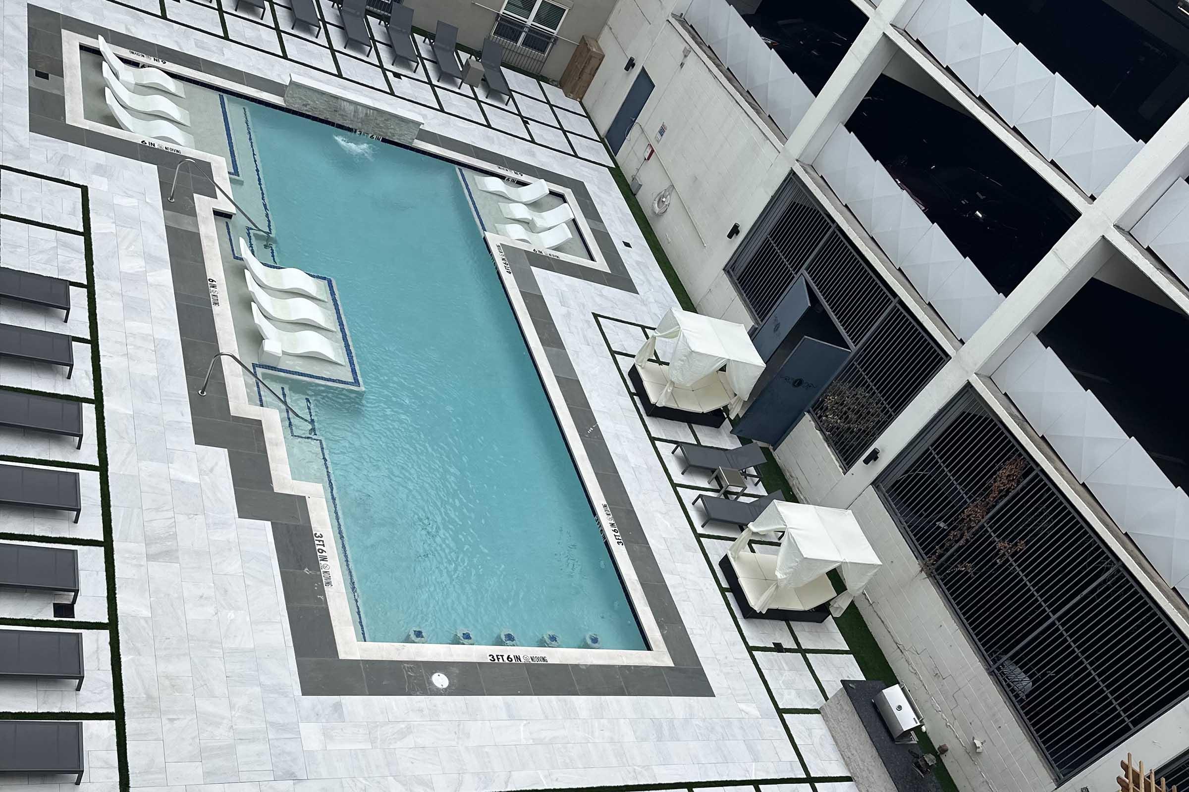 Aerial view of a modern swimming pool surrounded by decorative tiles and lounge chairs. The pool is rectangular with two curvy sections, featuring white lounge chairs on one side and cabanas on the other. The surrounding area is well-lit and includes structured landscaping.