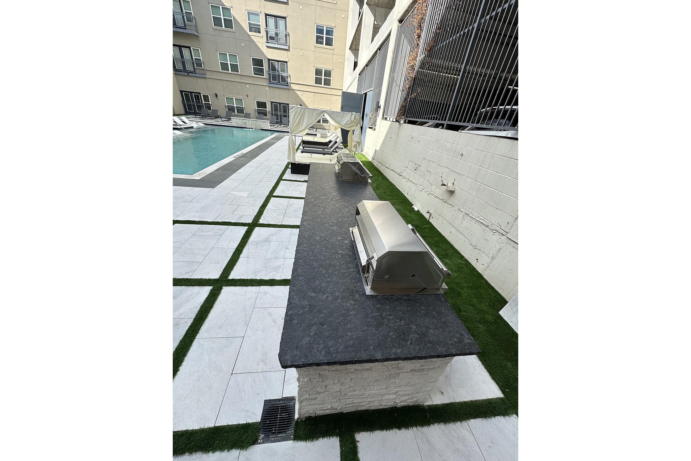 Outdoor space featuring a modern grilling area with two stainless steel grills on a black countertop. The setting includes a swimming pool in the background, surrounded by stone tiles and green grass patches, creating a stylish and inviting atmosphere for leisure and cooking.