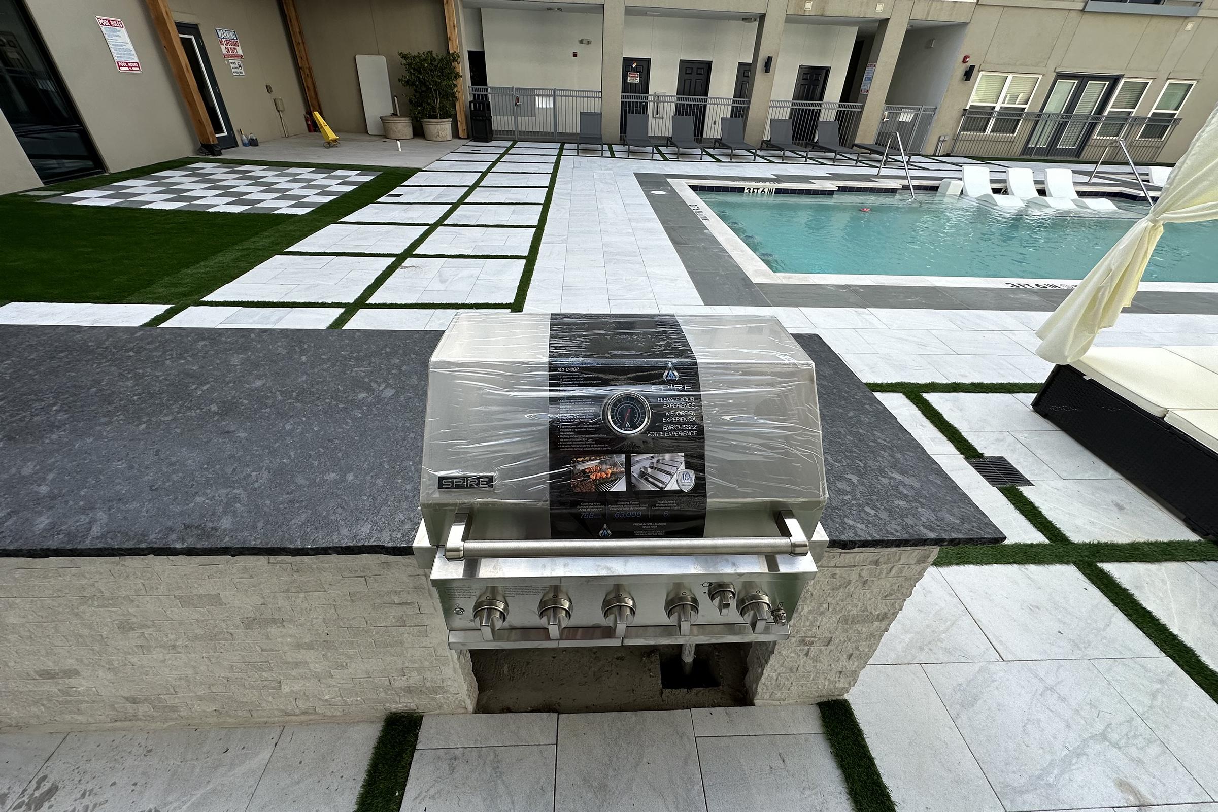 A stainless steel outdoor grill is positioned on a stone countertop, covered with plastic wrap. The grill is located in a modern pool area featuring a swimming pool, lounge chairs, and landscaped greenery. The surroundings include tiled flooring with a checkerboard pattern.