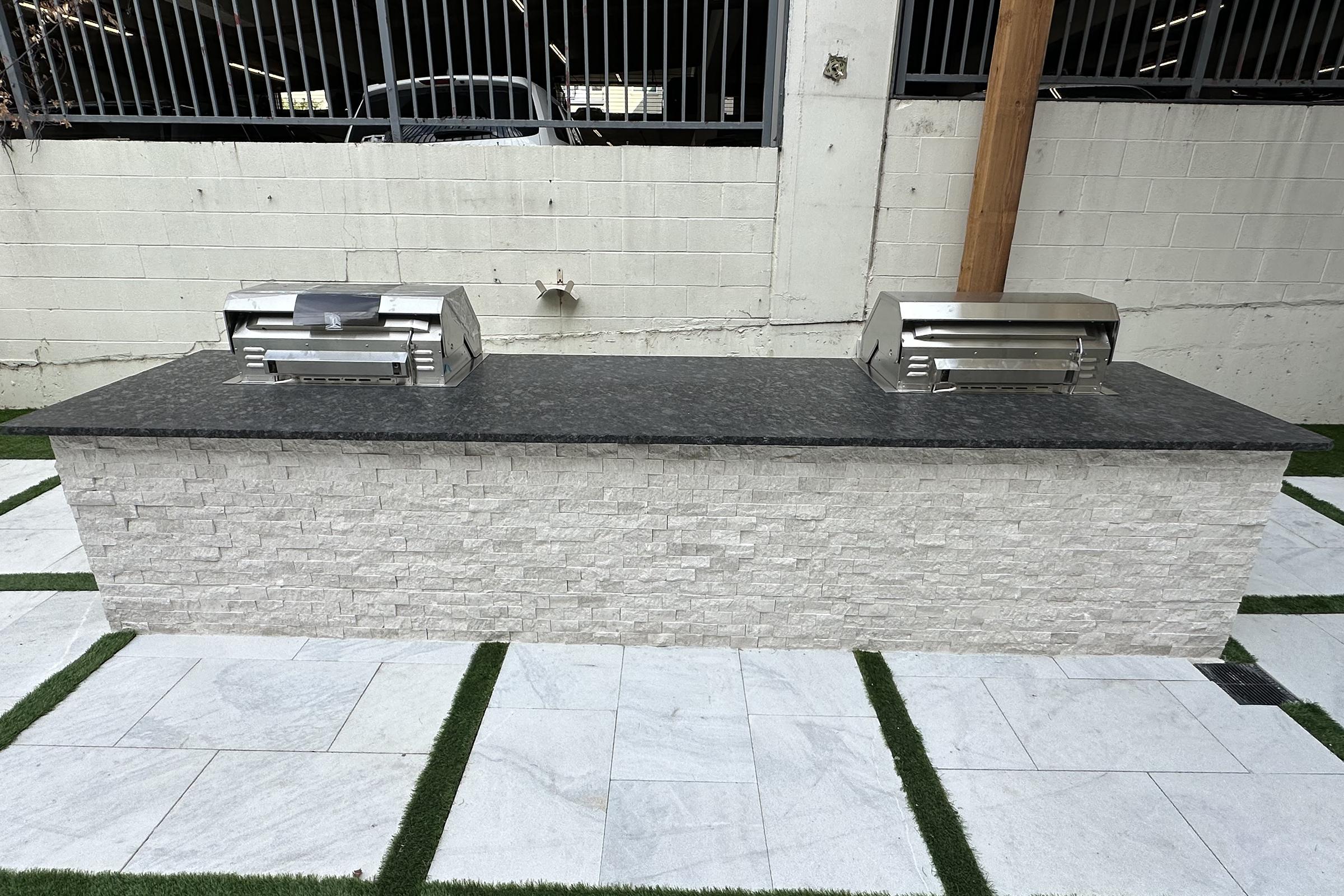 An outdoor grilling station featuring a stone countertop with two stainless steel grill units. The station is set against a white wall and surrounded by green grass and marble tiles, creating a modern and functional cooking space.