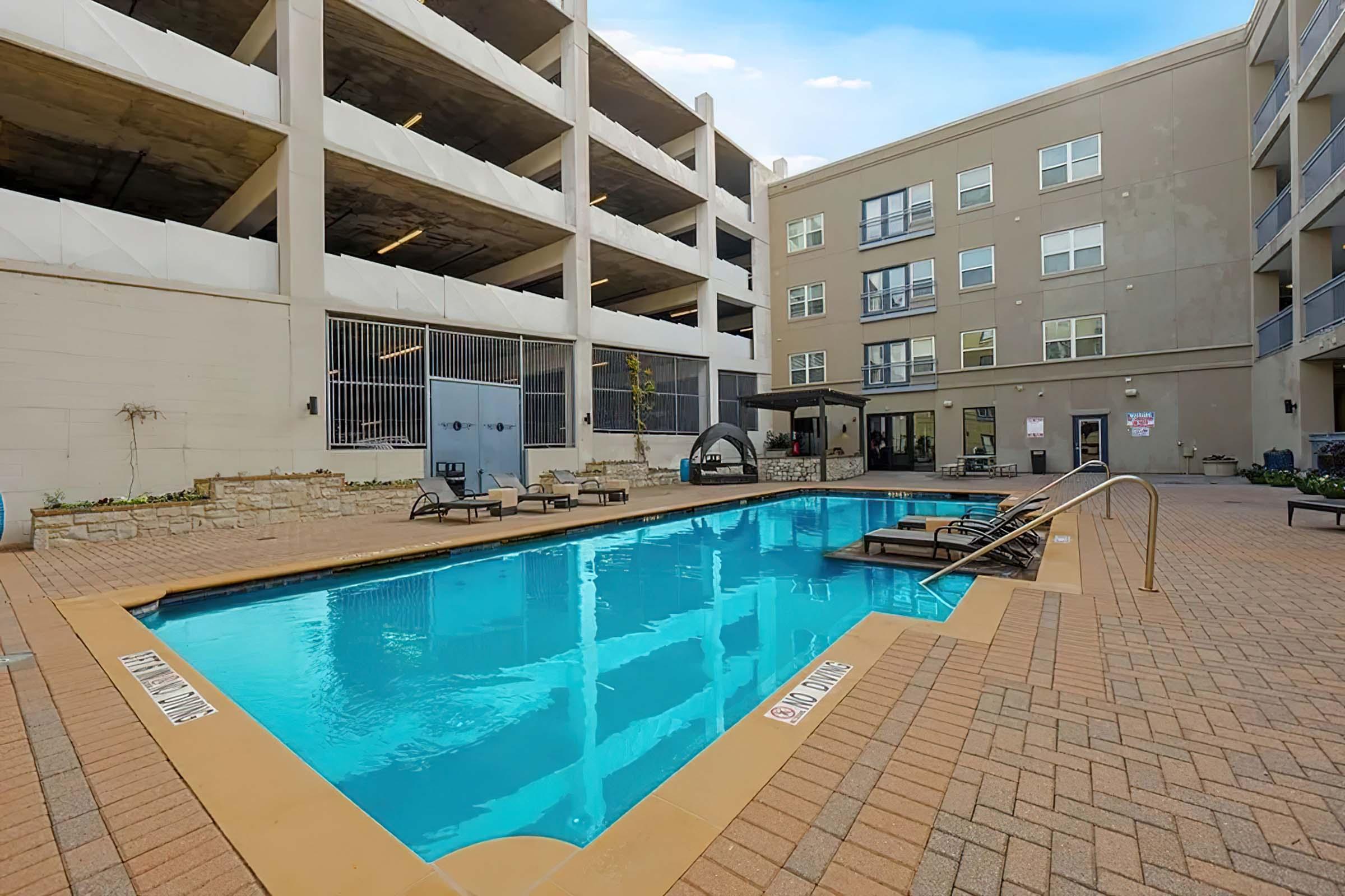 A spacious outdoor swimming pool surrounded by lounge chairs, with a modern apartment building and parking garage in the background. The area features well-maintained landscaping and a clear blue sky. Perfect for relaxation and leisure activities.