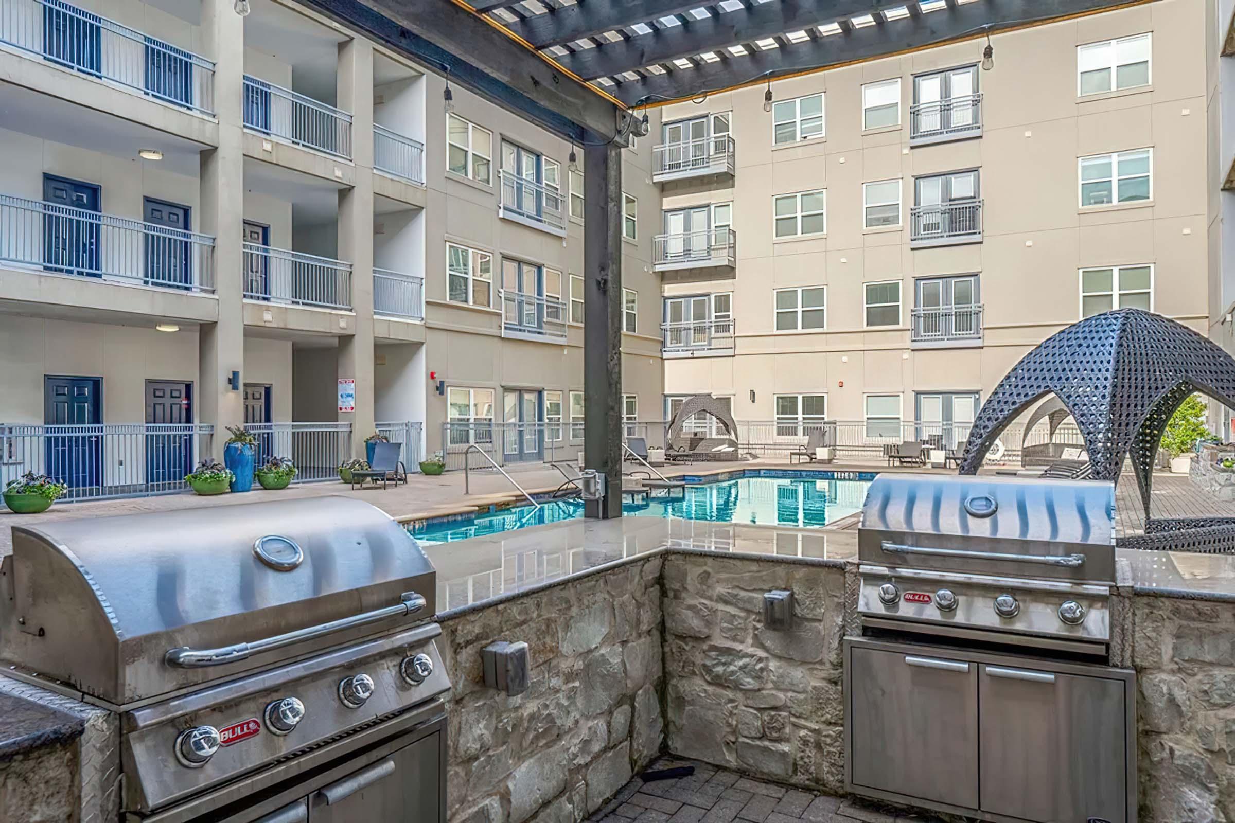 Outdoor area featuring two stainless steel barbecue grills beside a swimming pool. The pool is surrounded by lounge chairs and a stylish architectural structure overhead. Apartment buildings are visible in the background, creating a modern communal space.