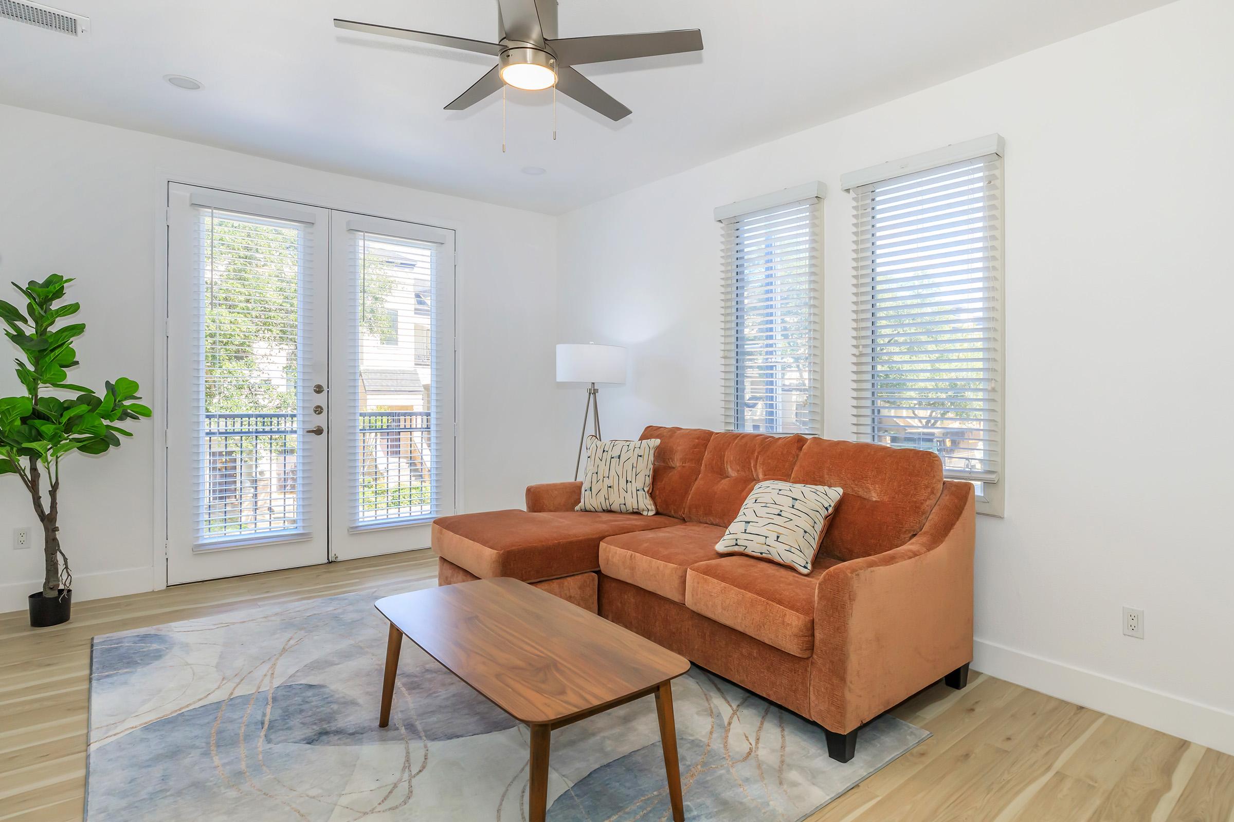 A stylish living room featuring a cozy orange sectional sofa with decorative pillows, a wooden coffee table, and a tall indoor plant. Large windows with blinds allow natural light, and a modern ceiling fan hangs overhead. The light wood flooring and neutral walls create a bright, inviting atmosphere.