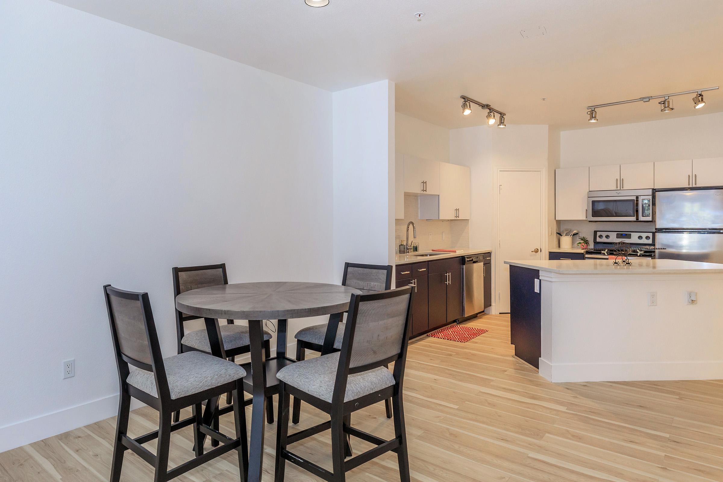 A bright, modern kitchen and dining area featuring a circular wooden table with four black chairs. The kitchen showcases stainless steel appliances, white cabinets, and wood flooring. Natural light floods the space, highlighting the clean, contemporary design.
