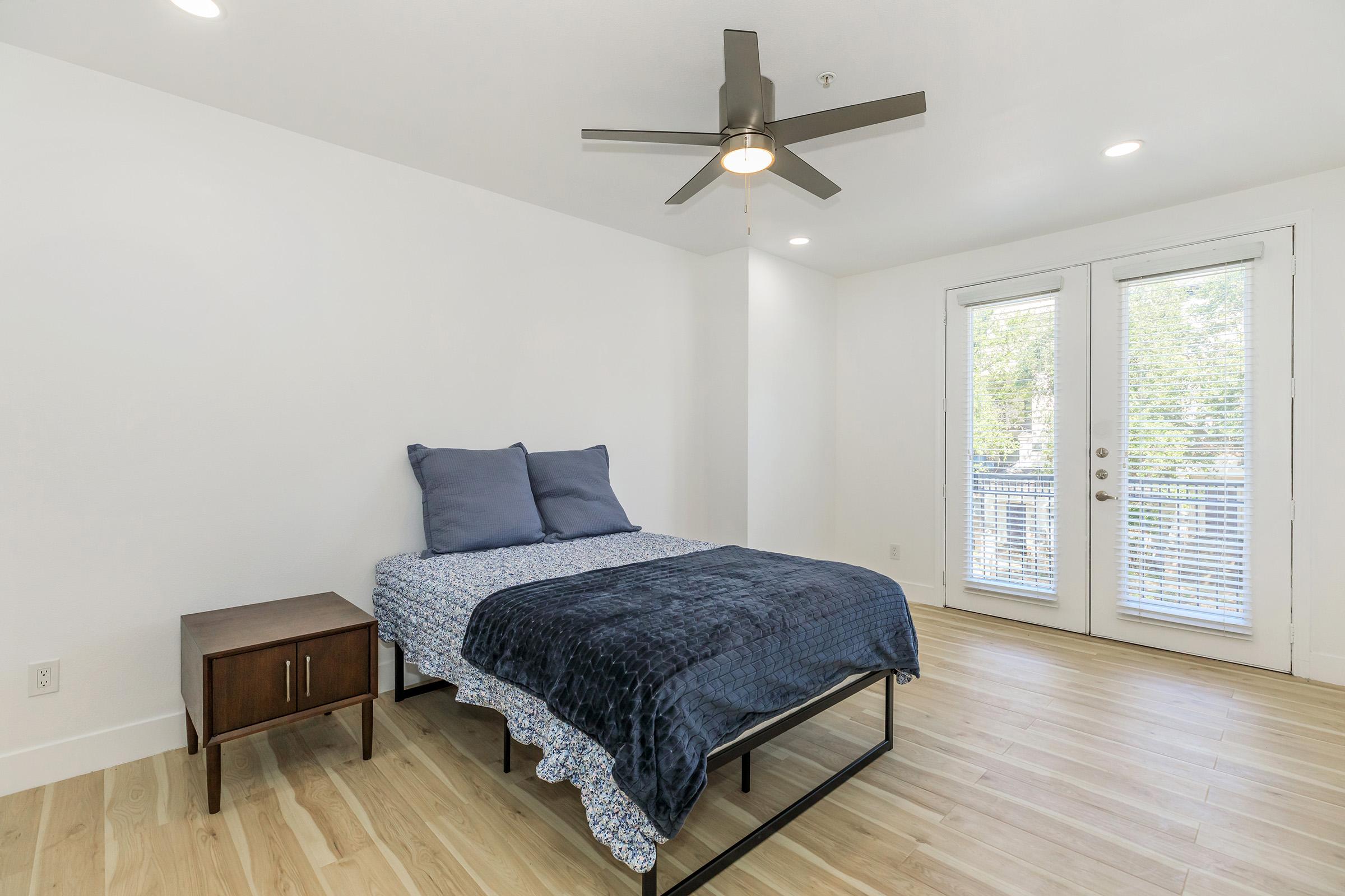 A bright, minimalistic bedroom featuring a bed with blue pillows and a dark blanket. A small wooden nightstand is beside the bed. Large sliding glass doors open to a balcony, letting in natural light. The wood floor adds warmth to the space, creating a cozy atmosphere.